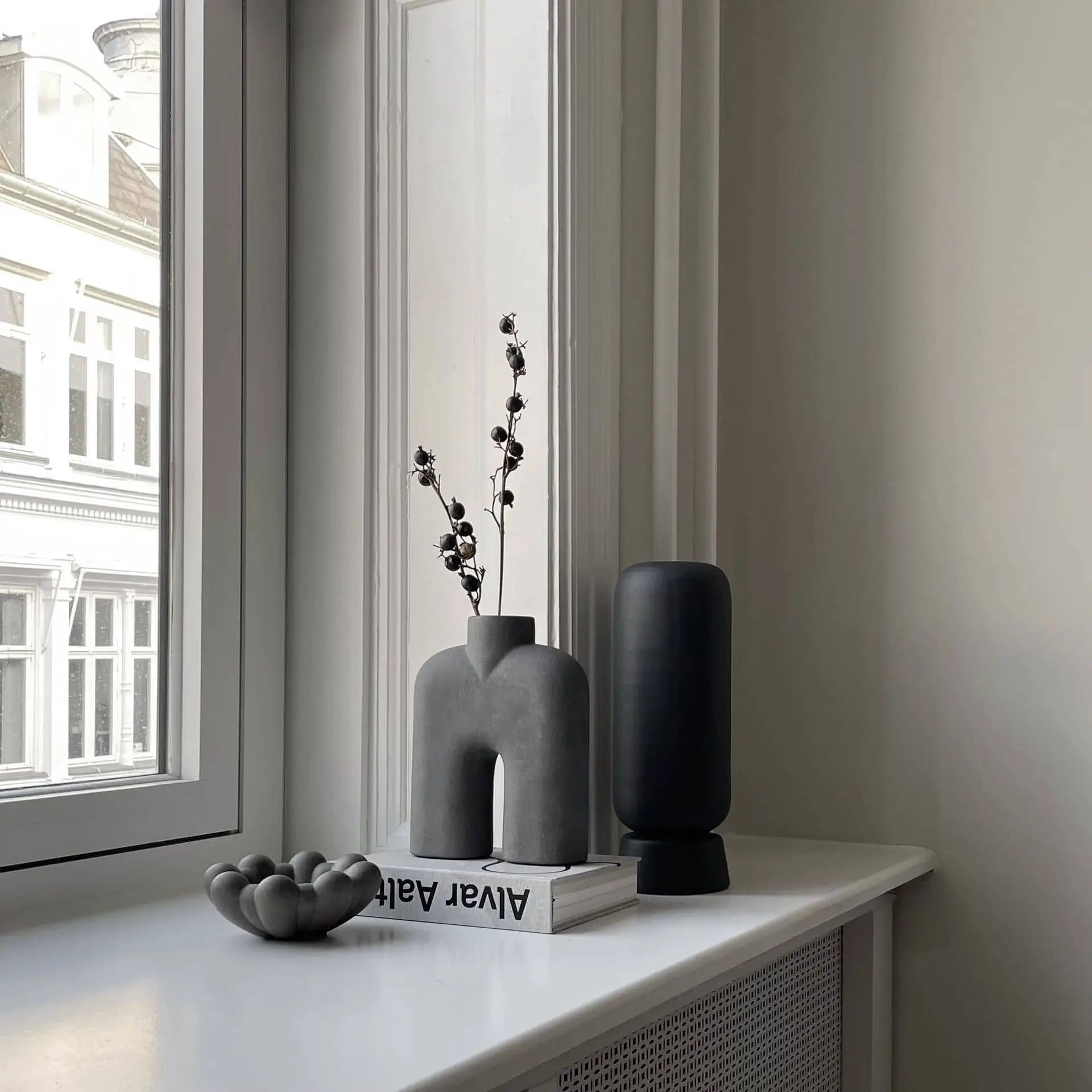 A minimalist windowsill displays a sculptural gray vase with dried branches, the 101 Copenhagen Bloom Tray, Mini, a stack of books, an organic-shaped sculpture, and a tall black vase, with buildings visible through the window.