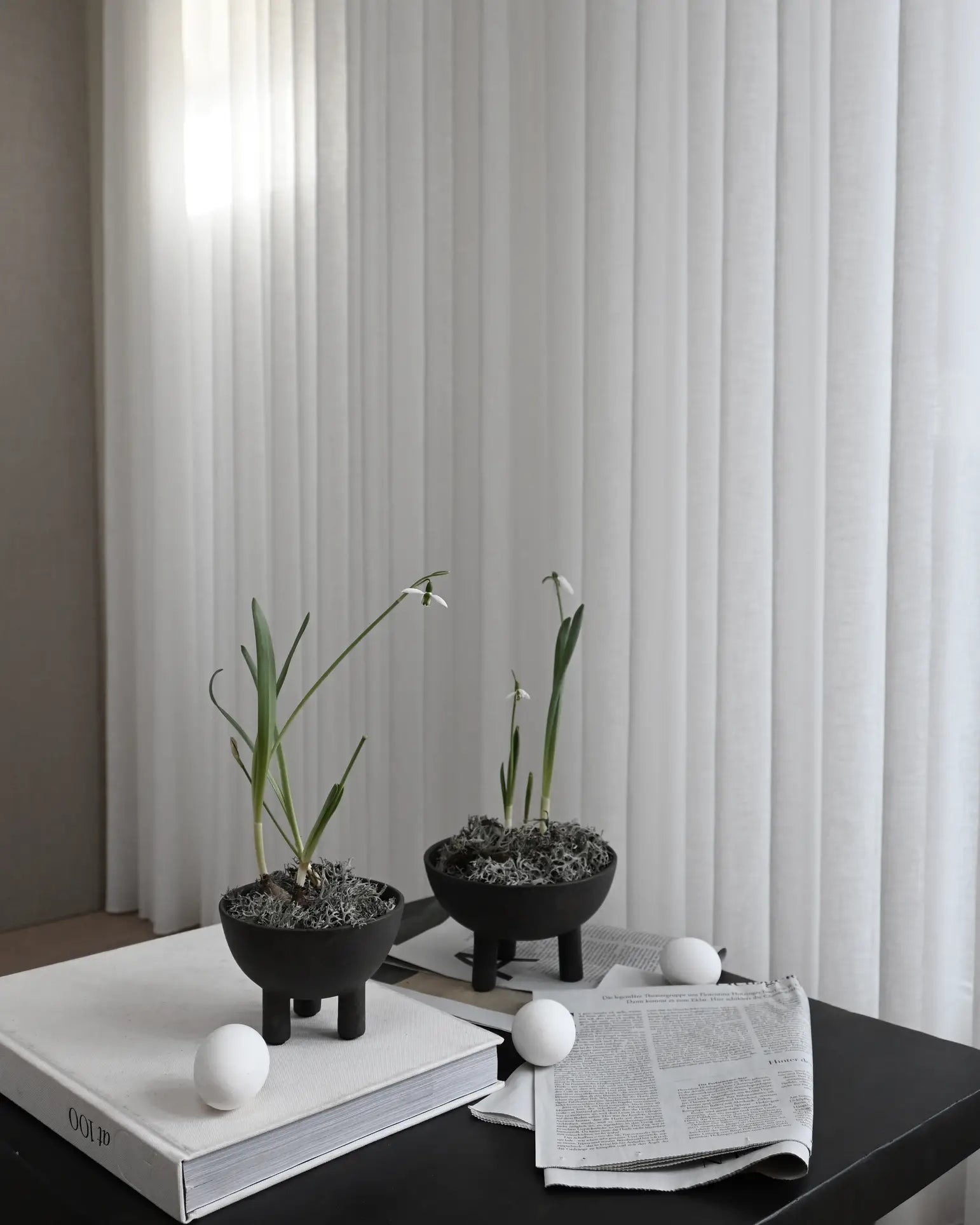 The 101 Copenhagen Duck Bowl, Big by 101 Copenhagen sits on a dark table beside white ping pong balls and open newspapers. Sheer white curtains in the background diffuse soft light, enhancing the Scandinavian-inspired scene.