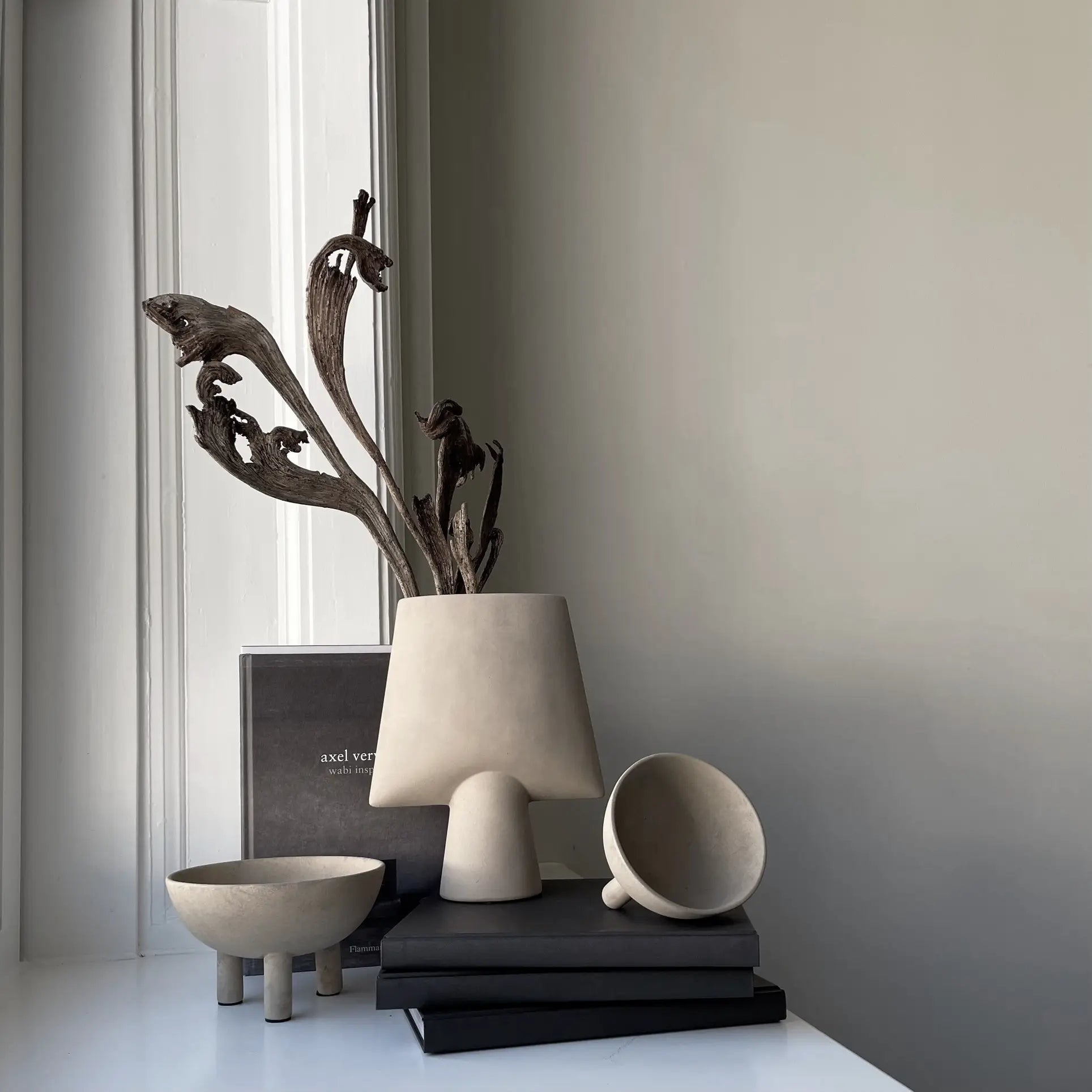 A minimalist scene with a 101 Copenhagen Duck Bowl, Big by 101 Copenhagen and dried branches, all displayed on stacked black books by a window with soft natural light and Scandinavian style.