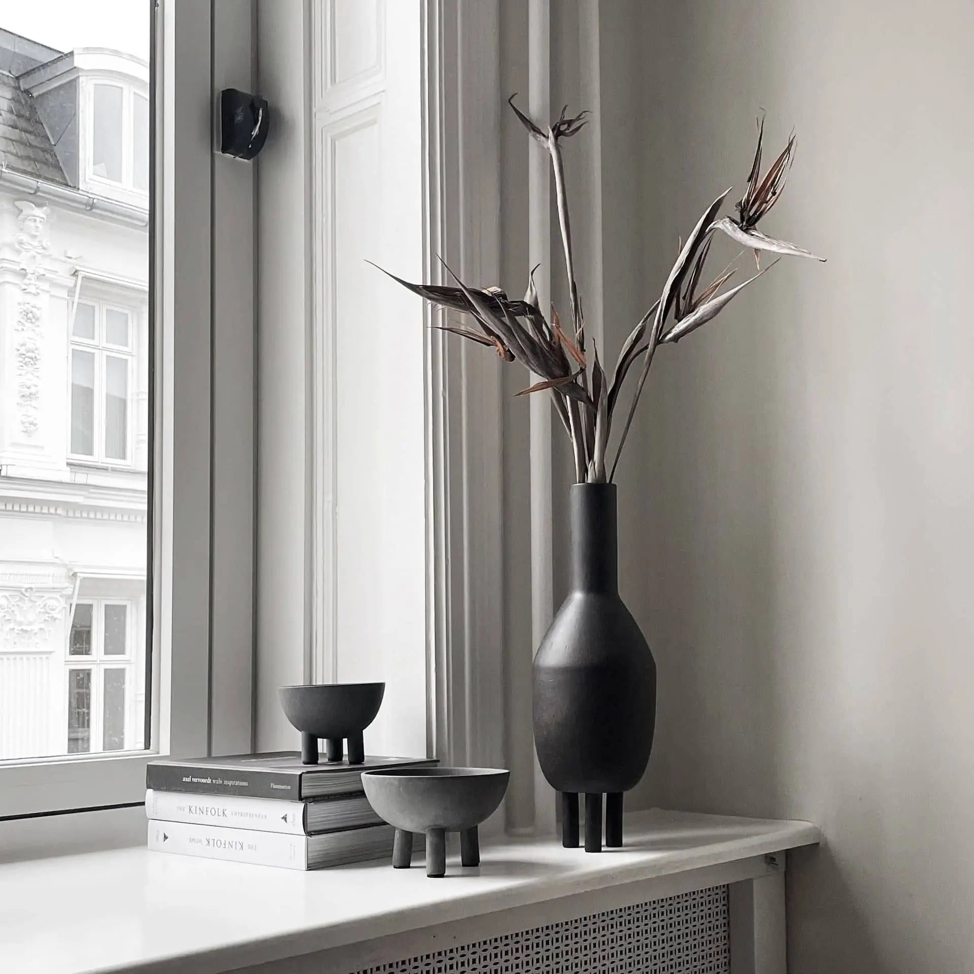 A minimalist interior features a black vase with dried branches, two 101 Copenhagen Duck Bowl Minis, and a stack of books on a white windowsill. Large windows fill the scene with natural light, enhancing the neutral home decor.