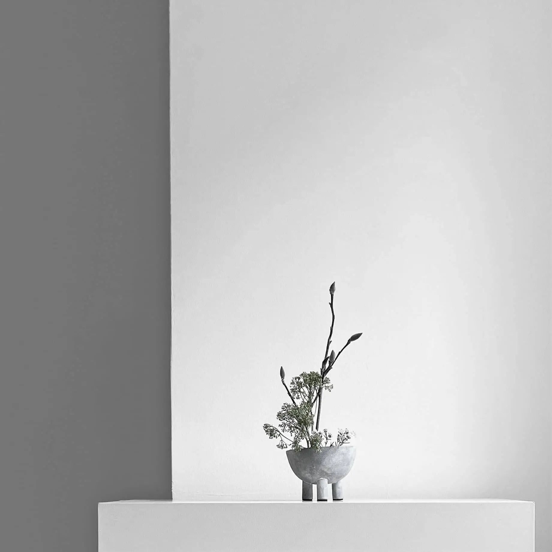 A minimalist scene features a small gray vase with delicate branches next to the 101 Copenhagen Duck Bowl, Mini, by 101 Copenhagen, elegantly displayed on a white surface against a light gray and white backdrop with clean lines.