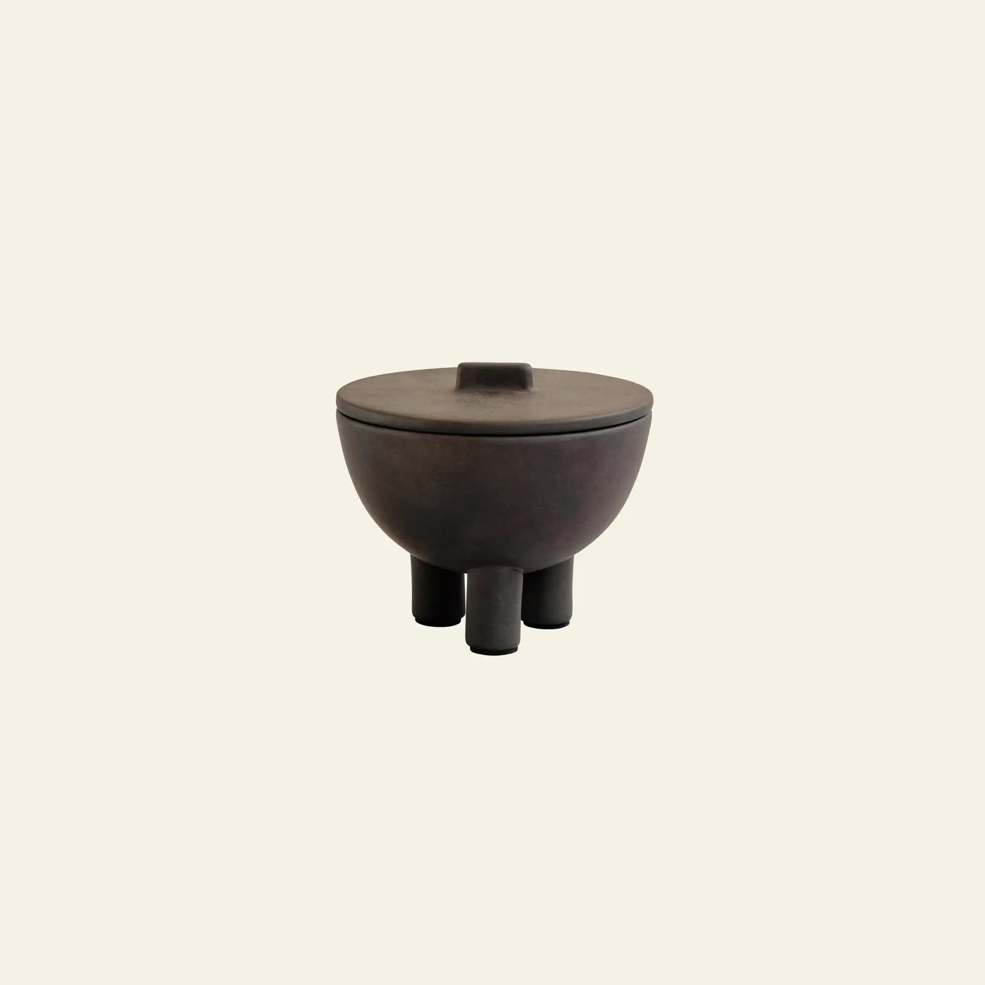 The 101 Copenhagen Duck Jar, Medio by 101 Copenhagen is a dark, round lidded jar with three cylindrical legs and a refined hand-glazed finish, offering unique decorative storage against a plain, light beige background.