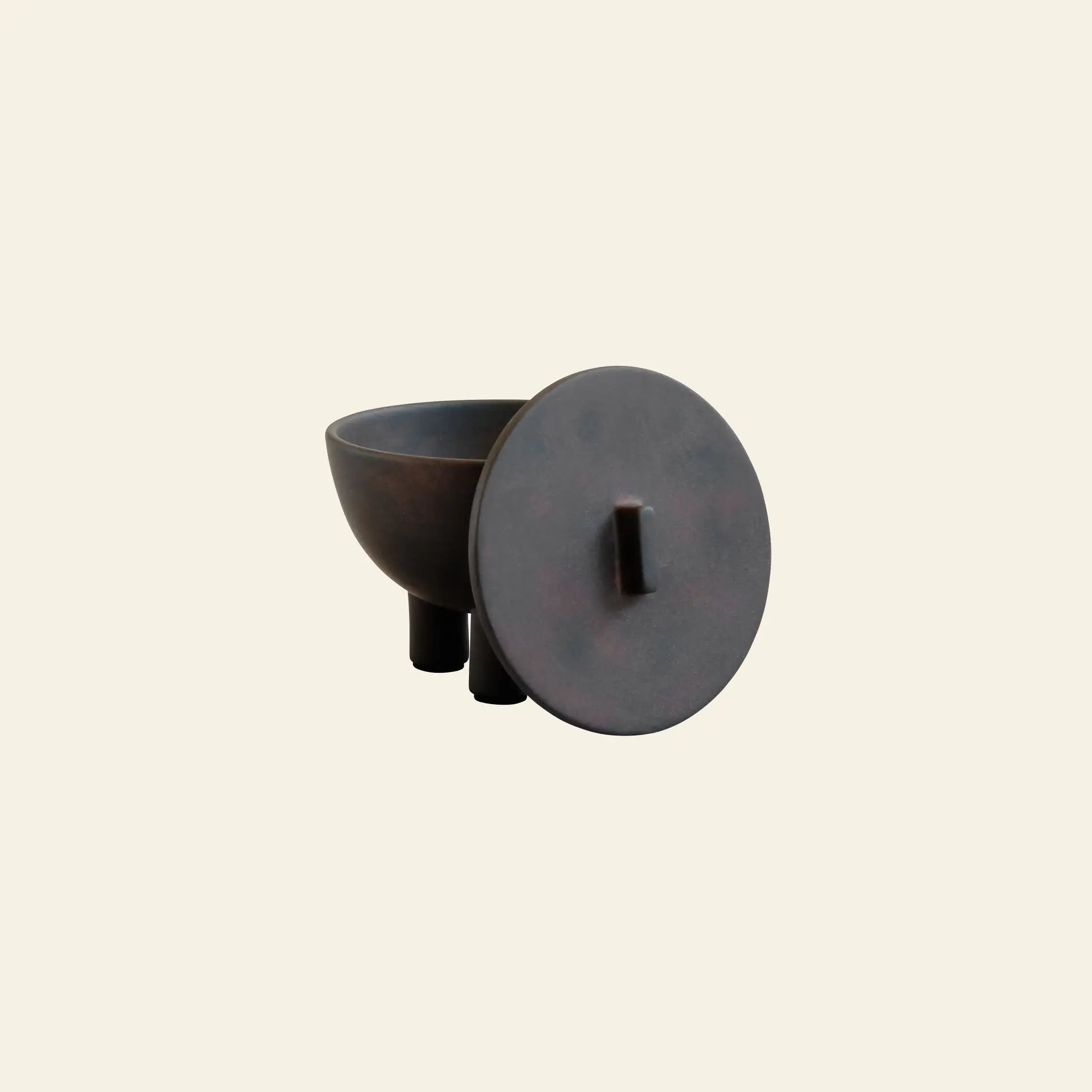 The 101 Copenhagen Duck Jar, Medio by 101 Copenhagen features a minimalist dark ceramic design with three legs and a round lid resting beside the bowl, hand-glazed for elegant decorative storage. Shown against a plain light background.