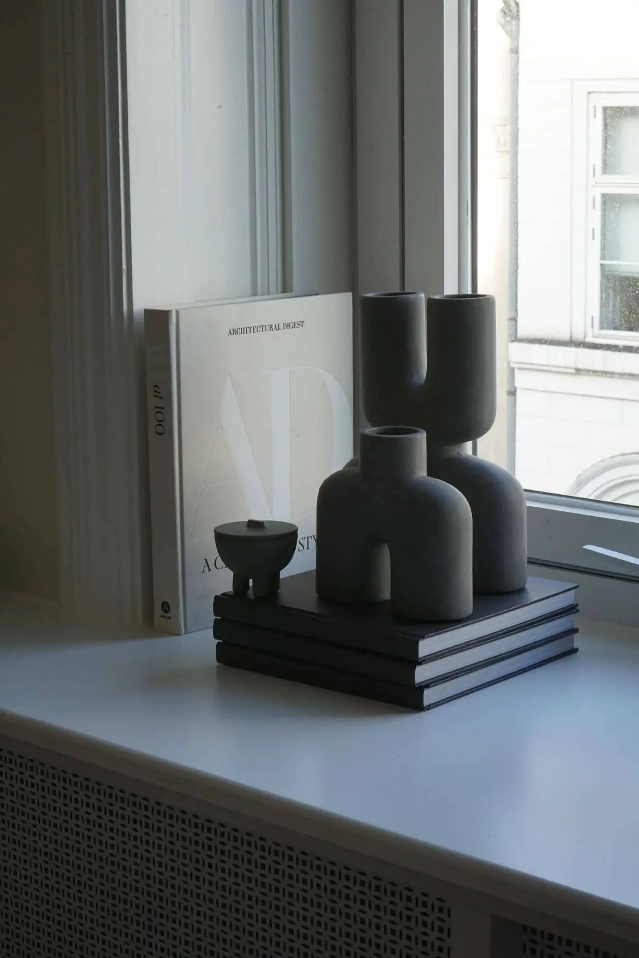 A minimalist windowsill features stacked books, sculptural gray vases, the 101 Copenhagen Duck Jar, Mini by 101 Copenhagen, a small bowl, and a large book titled Architectural Digest propped upright in natural light.