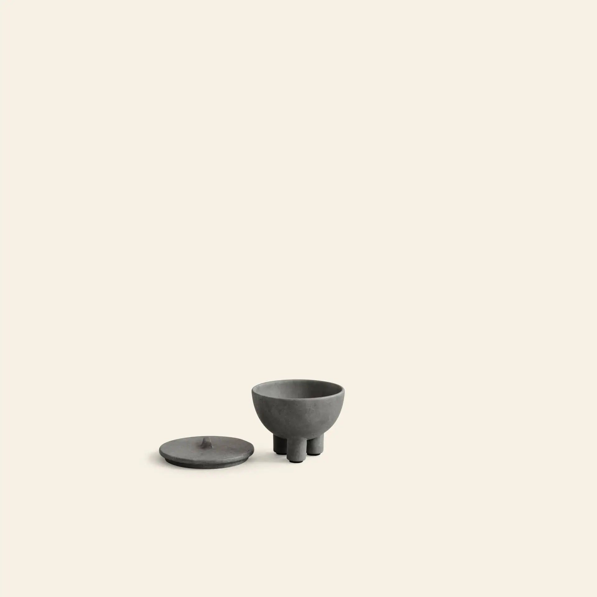 A minimalist image features the 101 Copenhagen Duck Jar, Mini by 101 Copenhagen in dark gray with three short legs and a matching flat lid beside it, set on a plain light beige background—an ideal decorative accent.