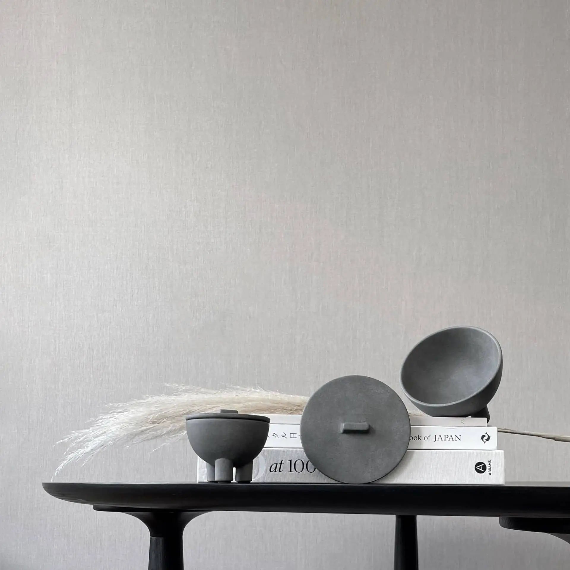 A minimalist tabletop features three modern black ceramic vessels, including the 101 Copenhagen Duck Jar, Mini by 101 Copenhagen, a stack of books, and dried grass sprigs, set against a plain light gray wall.