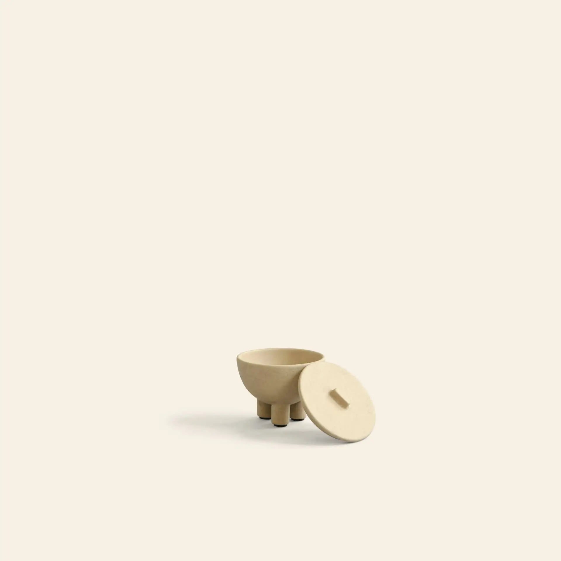 The 101 Copenhagen Duck Jar, Mini by 101 Copenhagen is a small beige storage jar with three short legs. Its matching lid, topped with a simple rectangular handle, rests beside it on an off-white background—perfect as a charming decorative piece.