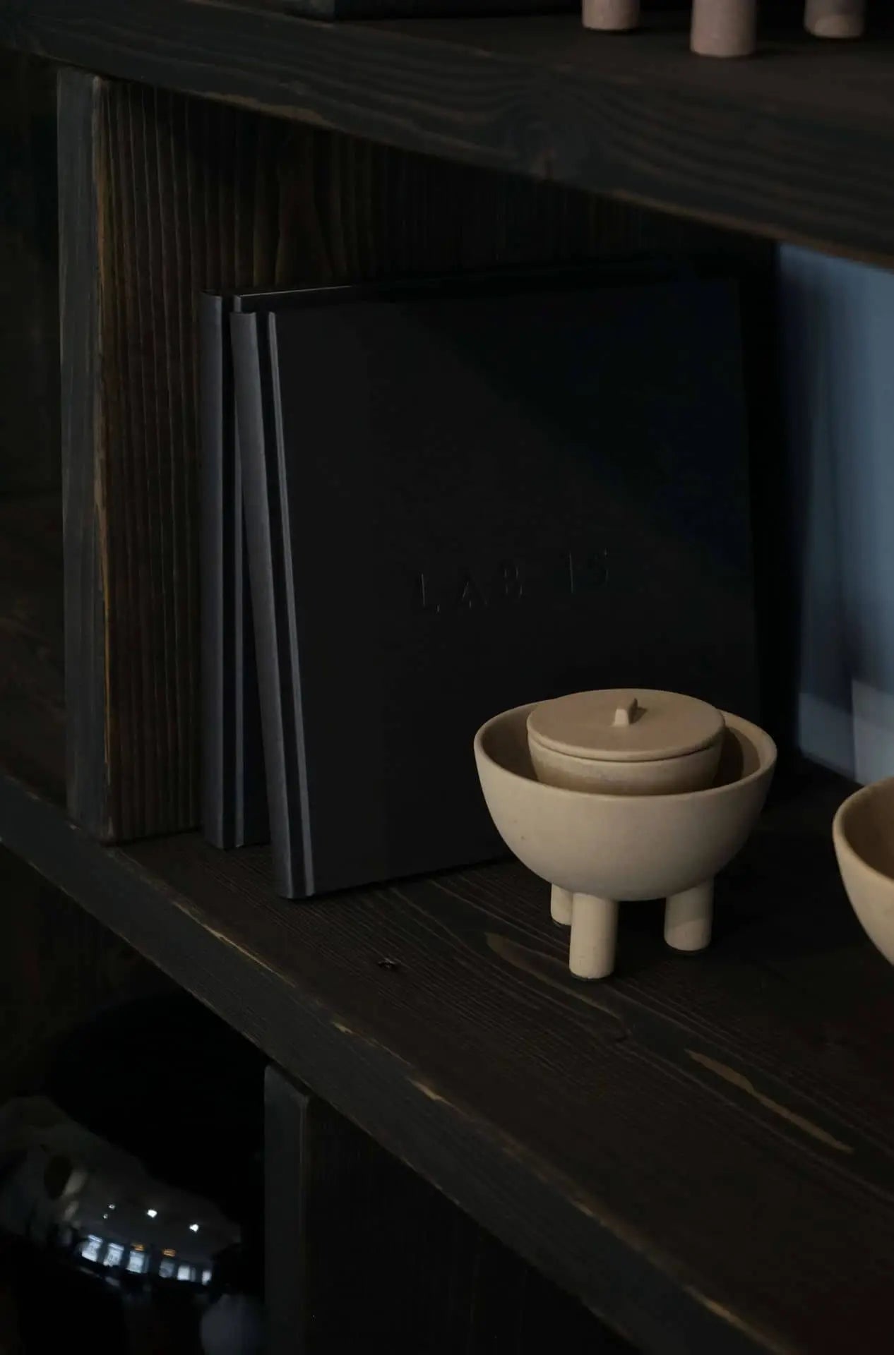 A dark wooden shelf holds two black upright books and the 101 Copenhagen Duck Jar, Mini—a small beige ceramic storage jar with lid and short legs. Another similar 101 Copenhagen piece is partially visible on the right.