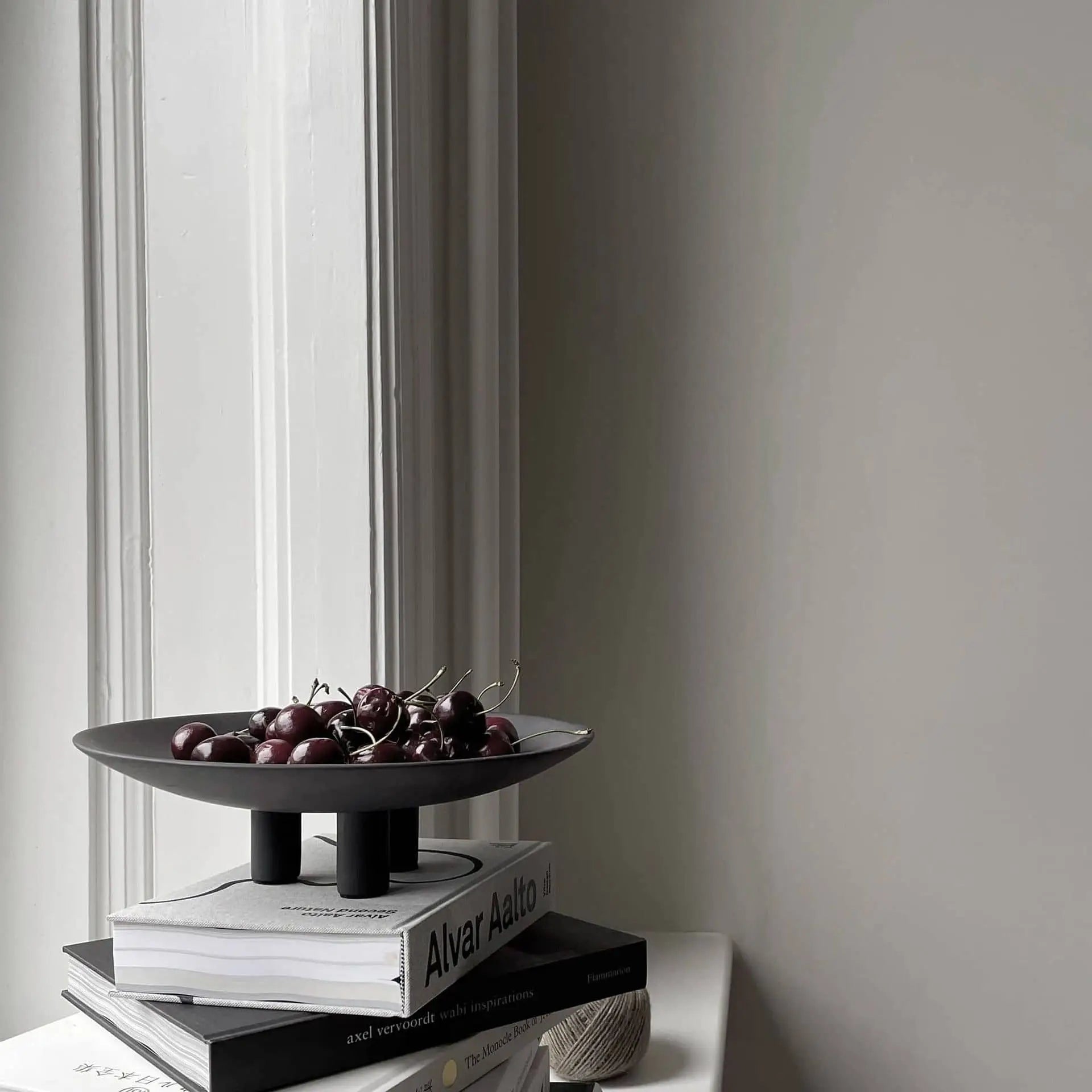 The 101 Copenhagen Duck Plate, Mini by 101 Copenhagen sits atop a stack of books near a bright window, filled with cherries and accompanied by a spool of twine—a minimalistic, softly lit accent for charming home decor.