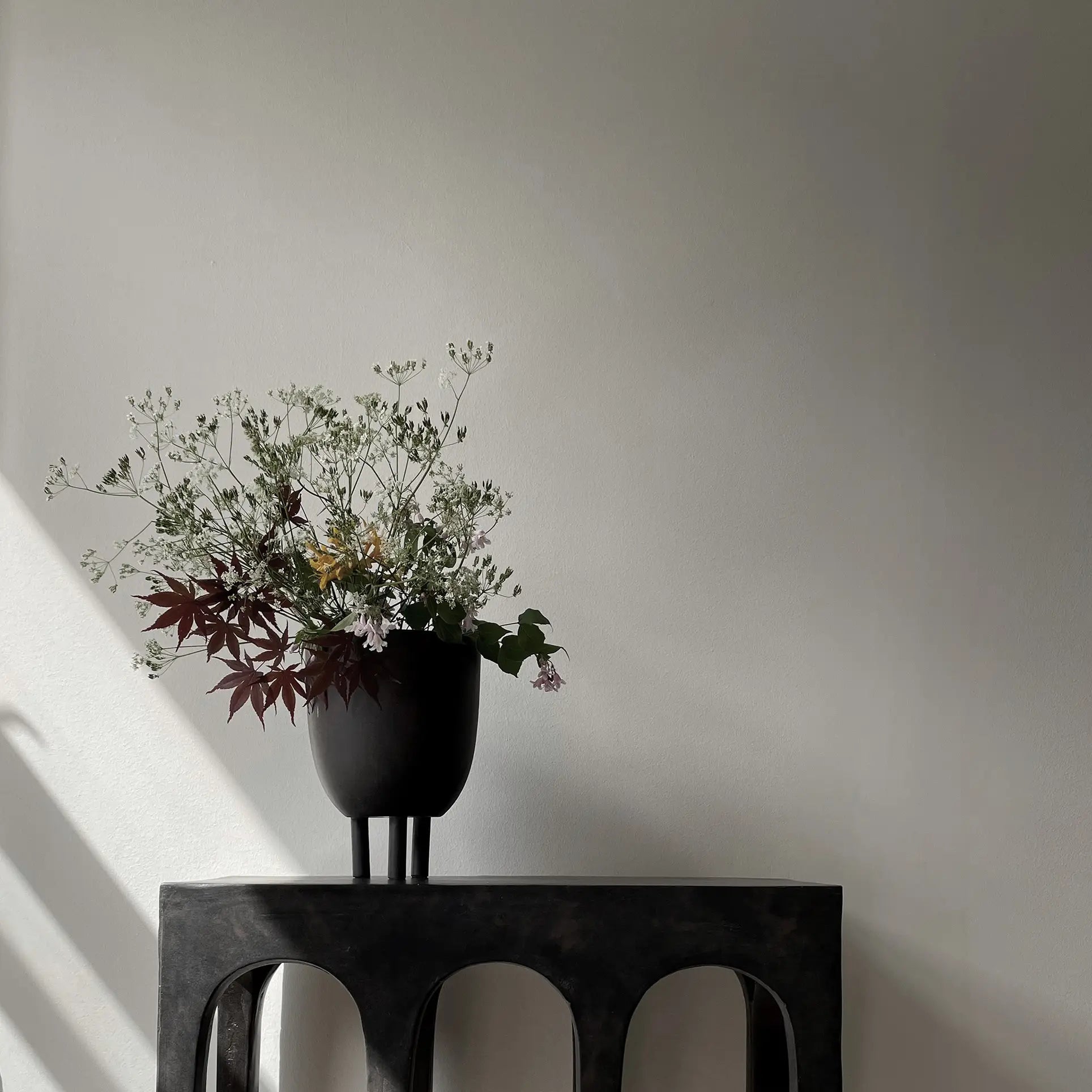 The 101 Copenhagen Duck Pot by 101 Copenhagen, filled with delicate white and burgundy flowers, sits on a dark arched table, adding whimsical charm against a light gray wall where sunlight casts elegant shadows.
