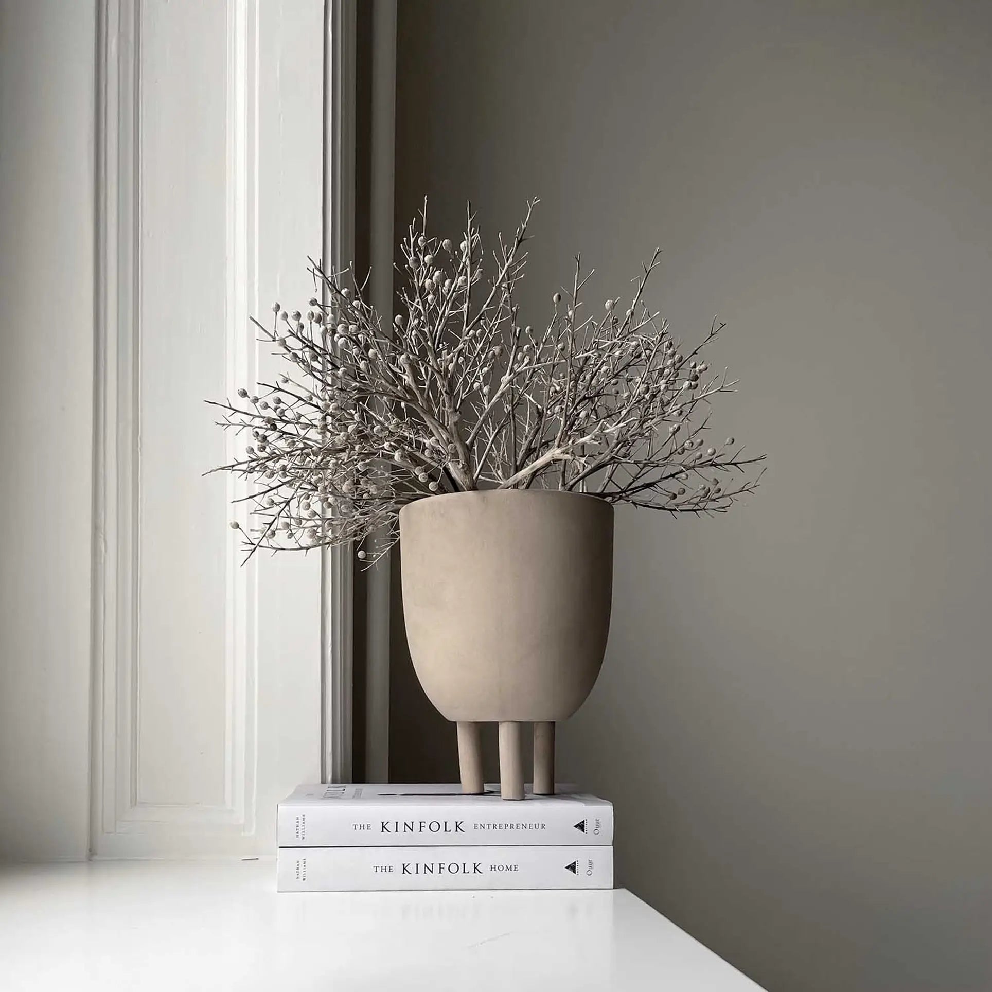 A minimalist setup features the 101 Copenhagen Duck Pot by 101 Copenhagen with dried branches, displayed on two stacked books—The Kinfolk Home and The Kinfolk Entrepreneur—highlighting Scandinavian design near a light wall and window.