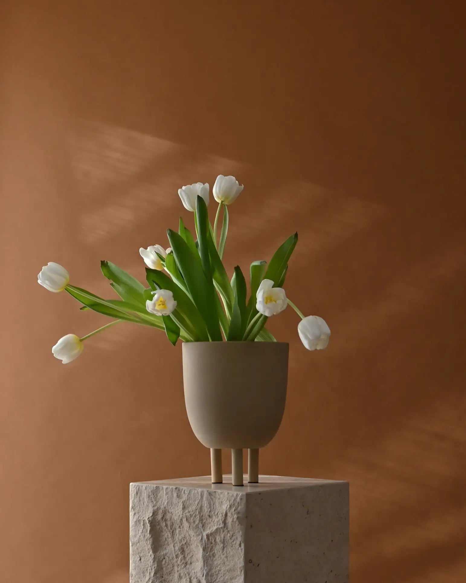 The 101 Copenhagen Duck Pot vase by 101 Copenhagen, featuring three short legs and a beige finish, displays white tulips with green leaves on a textured stone pedestal against a brown backdrop, bringing whimsical charm to minimalist décor.