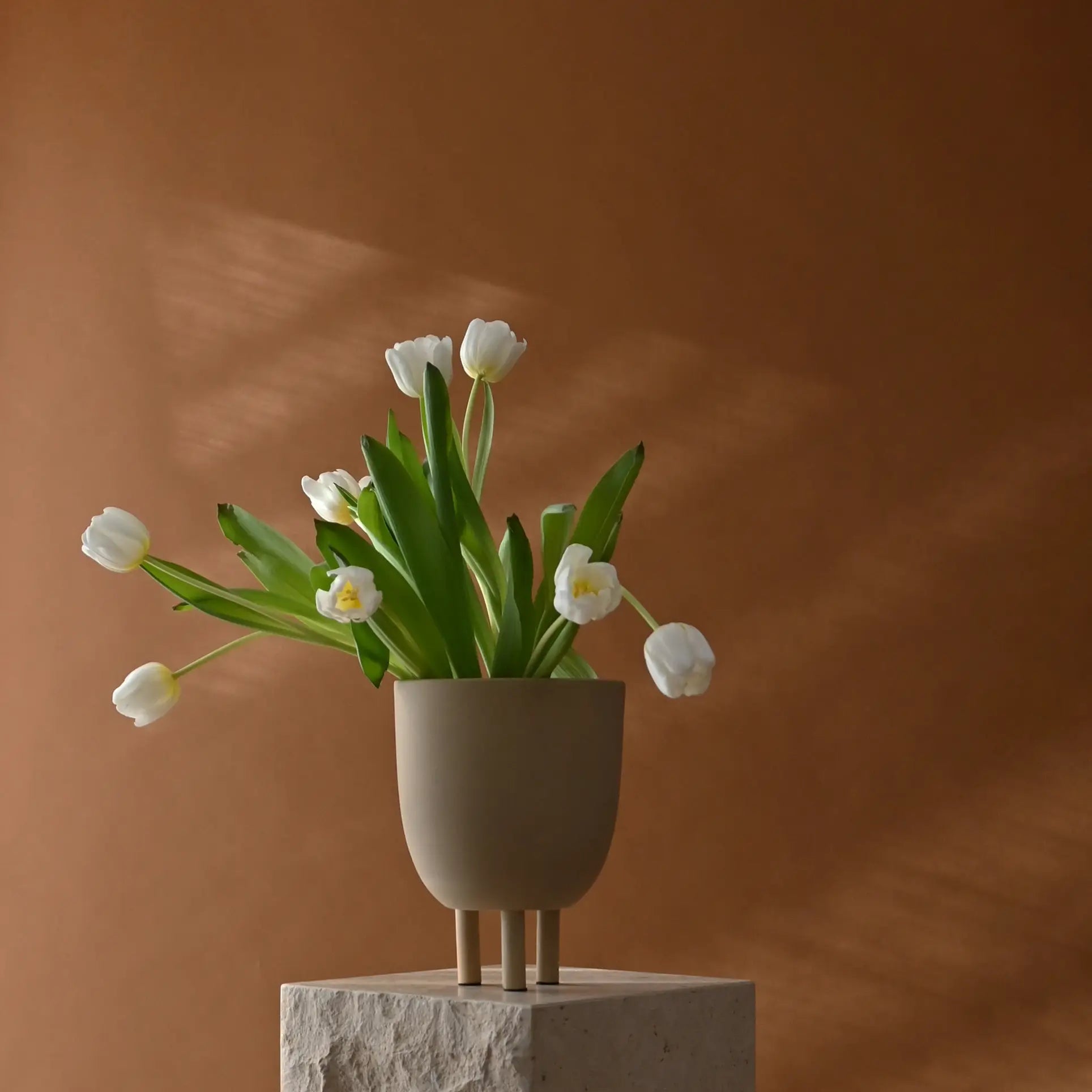 The 101 Copenhagen Duck Pot by 101 Copenhagen, a beige three-legged vase inspired by Scandinavian design, holds white tulips with green leaves on a textured stone pedestal with a plain brown backdrop and soft lighting.