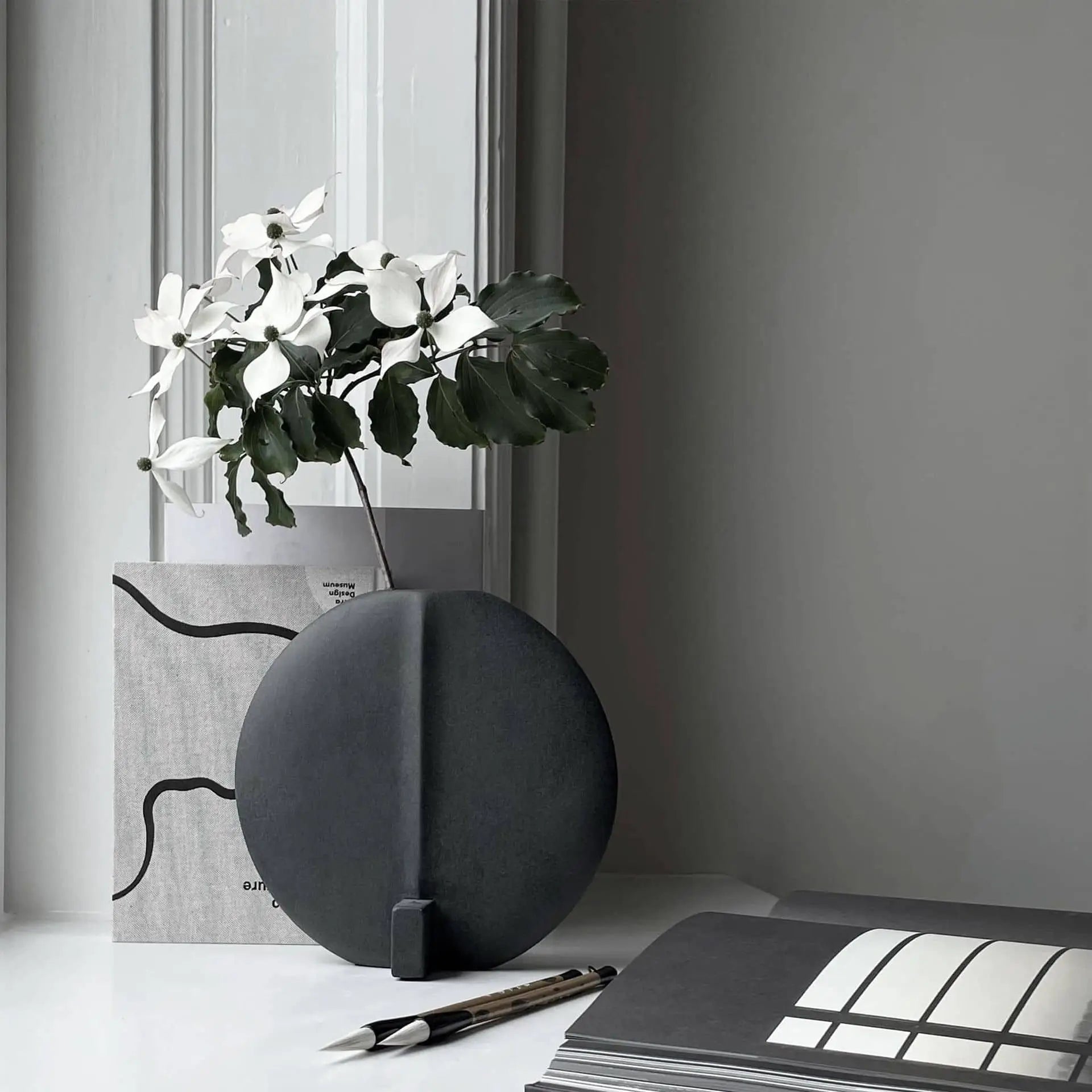 A minimalist scene features the 101 Copenhagen Guggenheim Vase, Petit—a round black, hand-glazed piece—holding white flowers with green leaves on a white surface, beside a pen, open book, and abstract art set against a gray and white background.