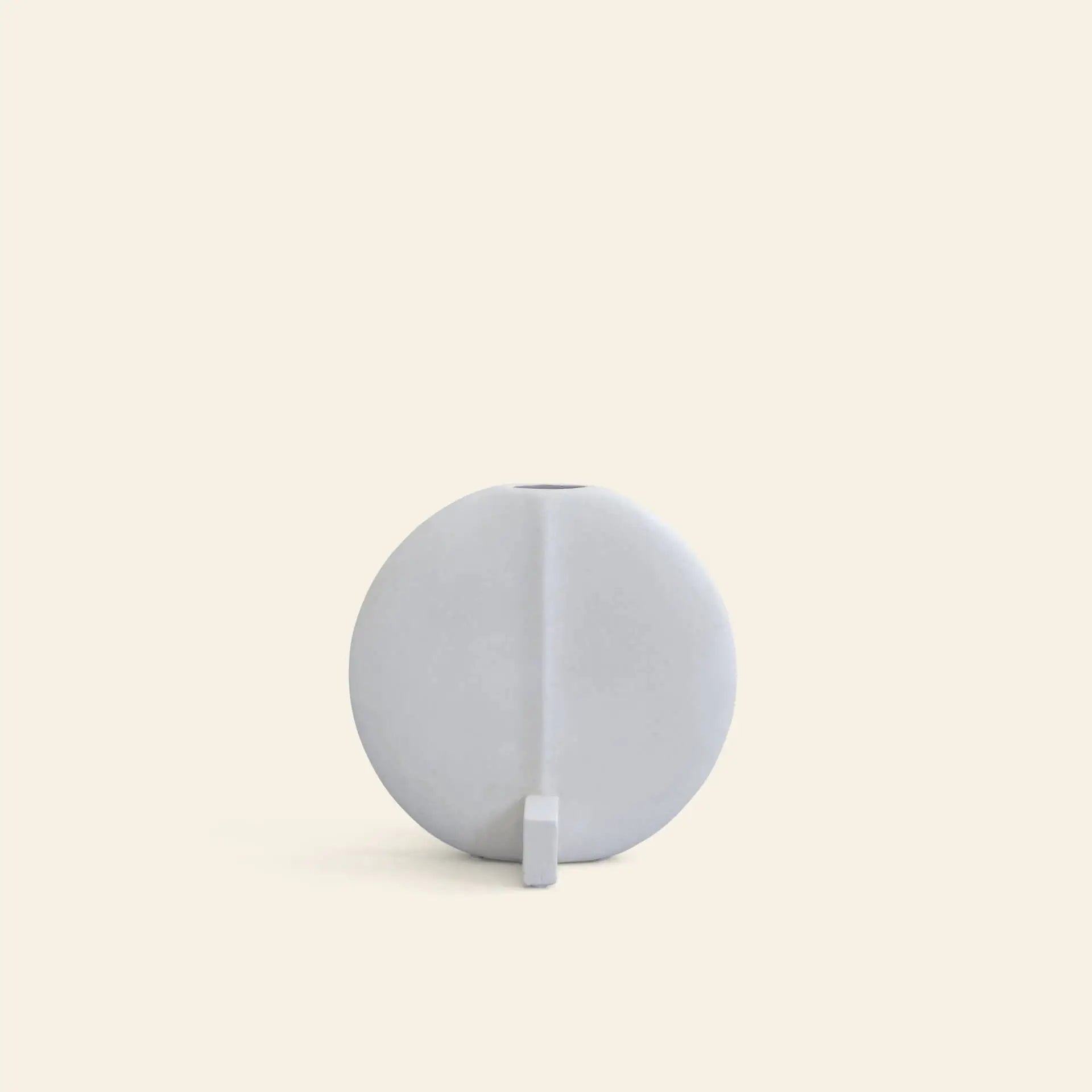 The 101 Copenhagen Guggenheim Vase, Petit by 101 Copenhagen is a minimalist round white ceramic piece with a matte hand-glazed finish and small rectangular base, evoking subtle art deco flair against a plain beige background.
