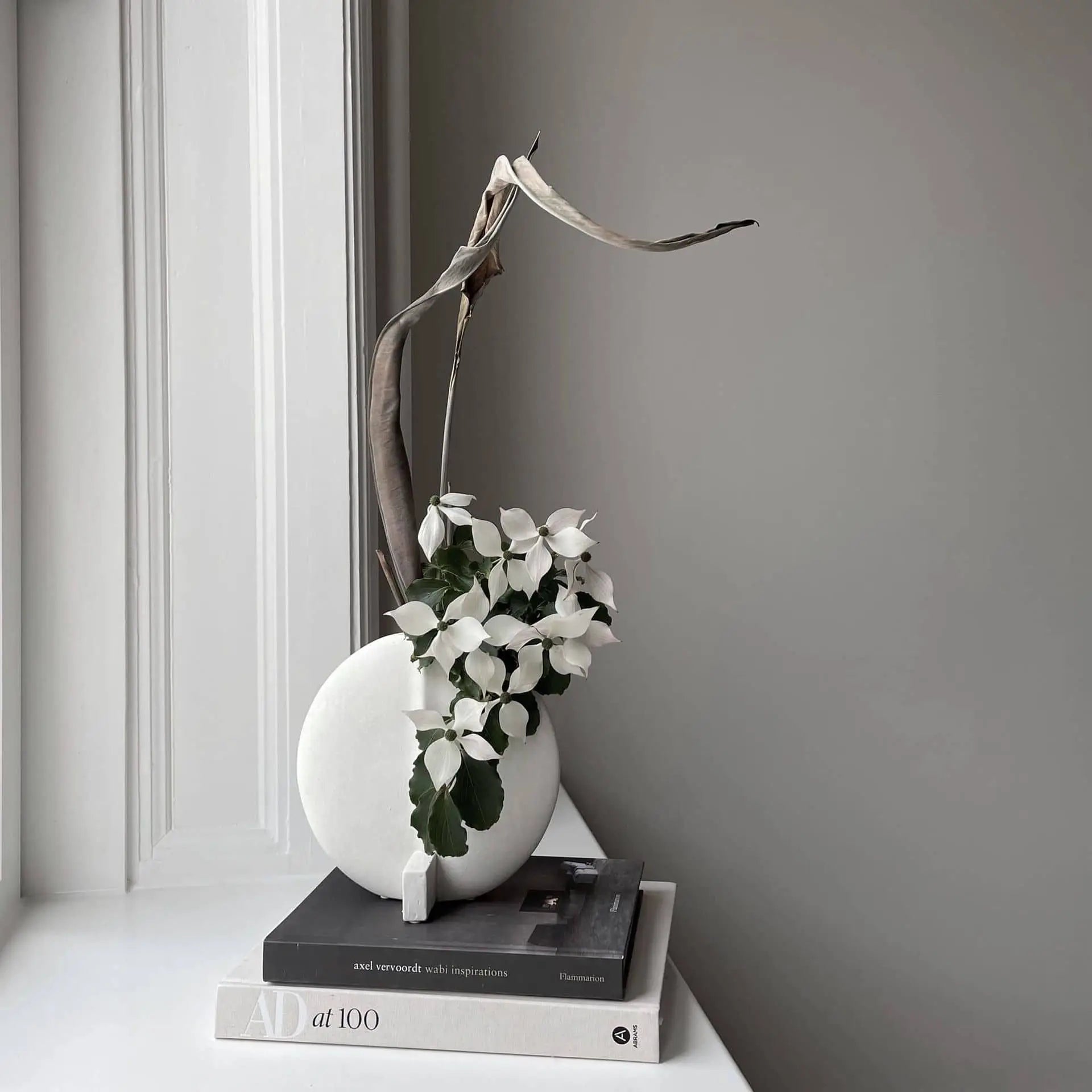 The 101 Copenhagen Guggenheim Vase, Petit by 101 Copenhagen, hand-glazed in white and styled with a dried leaf and white flowers, rests on two monochrome books by a window for a calm, modern Art Deco-inspired look.