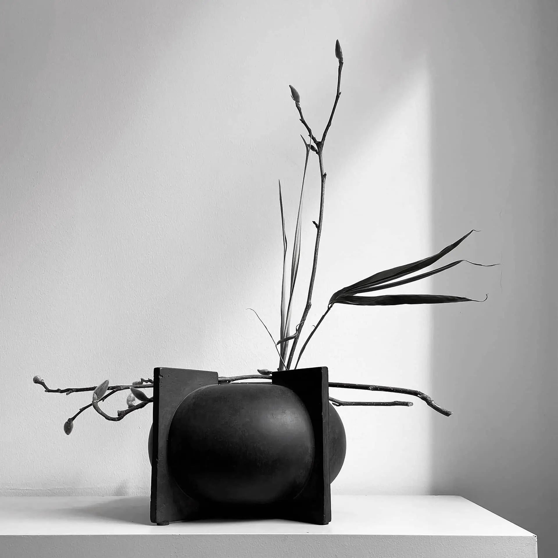 The 101 Copenhagen Kyoto Vase, Mini by 101 Copenhagen features a minimalist black ceramic design with geometric sides, displayed with bare branches and long leaves on a white surface against a plain wall with dramatic lighting.