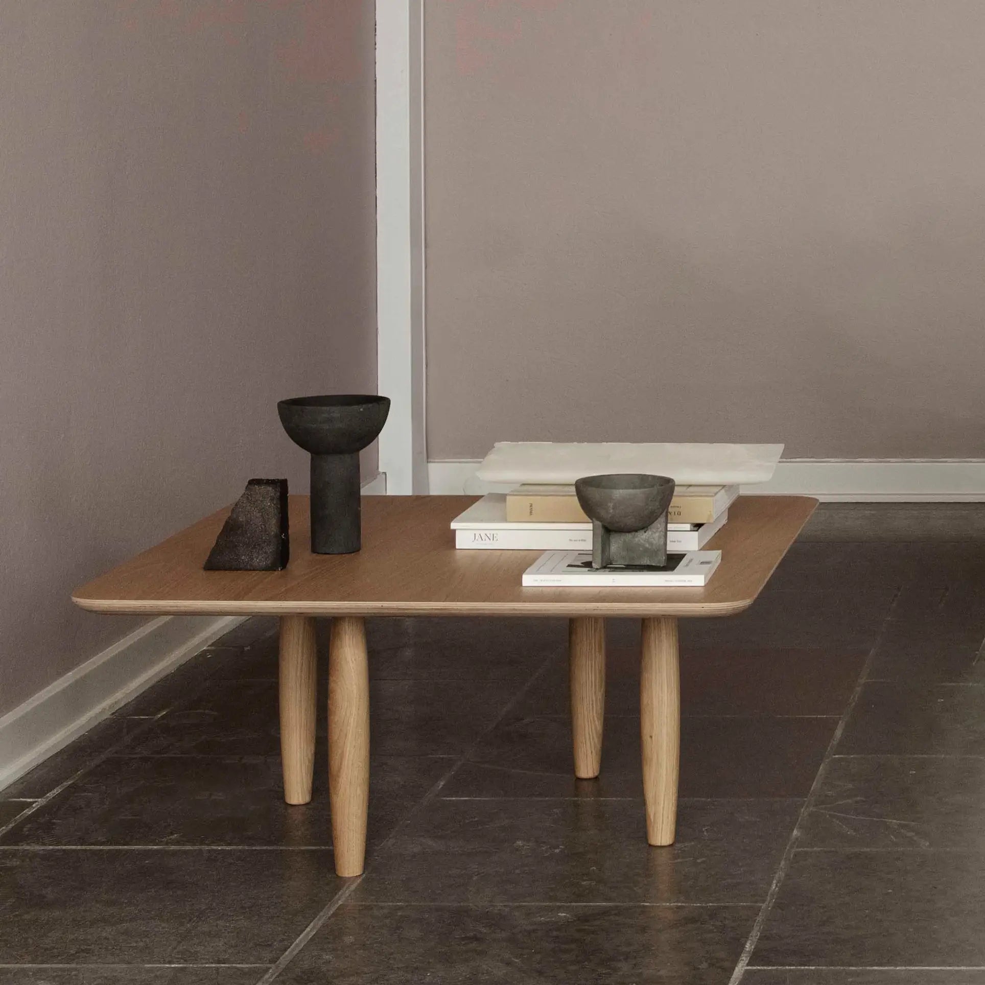 A minimalist wooden coffee table with rounded legs displays 101 Copenhagen Osaka Bowl, Mini, a stone sculpture, and stacked books on a tiled floor next to a mauve wall.