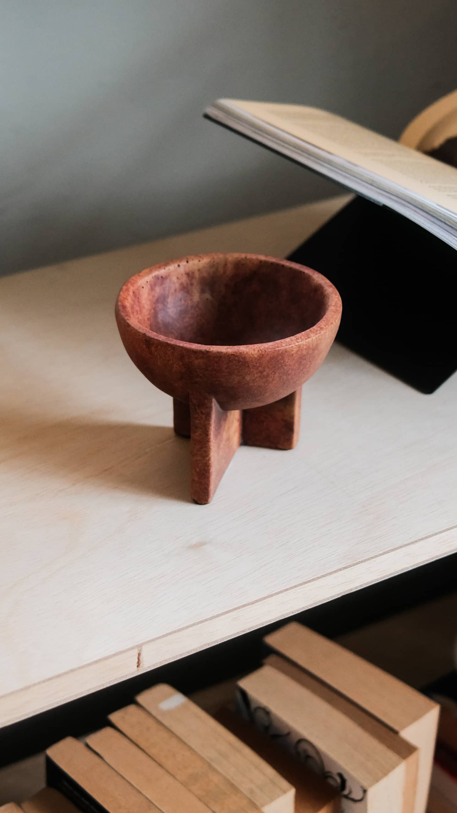 The 101 Copenhagen Osaka Bowl, Mini by 101 Copenhagen is a small, round, reddish-brown ceramic bowl with three sturdy legs, sitting on a light wooden surface near books and echoing the elegant Osaka bowl style.