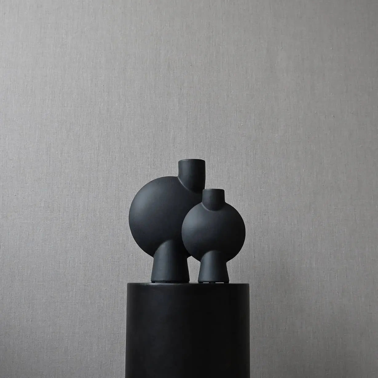 Two 101 Copenhagen Sphere Vase Bubl, Mini vases by 101 Copenhagen in matte black with geometric round bodies and small necks rest on a cylindrical pedestal, creating striking décor against a light gray textured background.