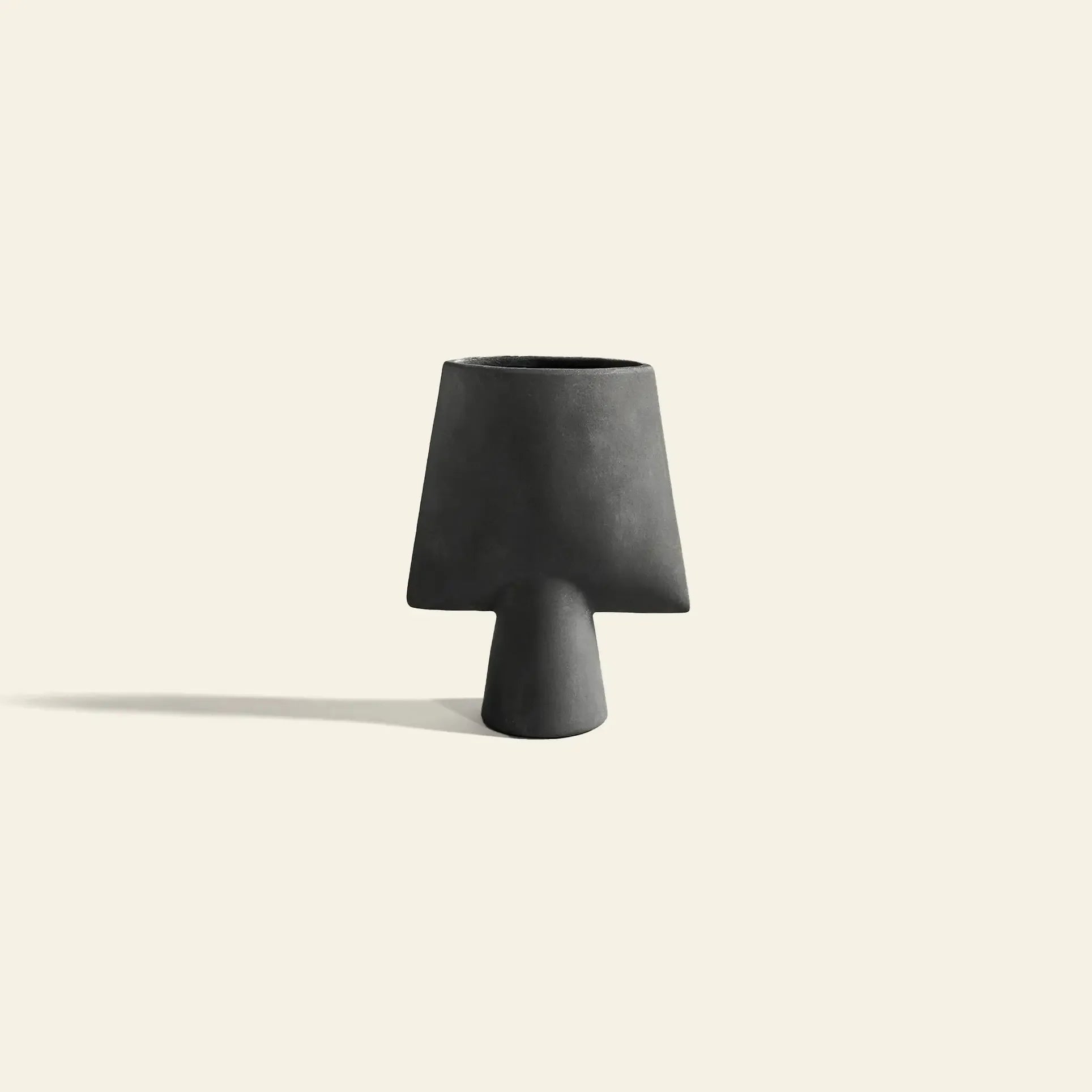 The 101 Copenhagen Sphere Vase Square, Mini by 101 Copenhagen features a matte black ceramic finish with a wide, flared top and narrow base, evoking ancient sculptural elegance on a light beige background with a soft left shadow.