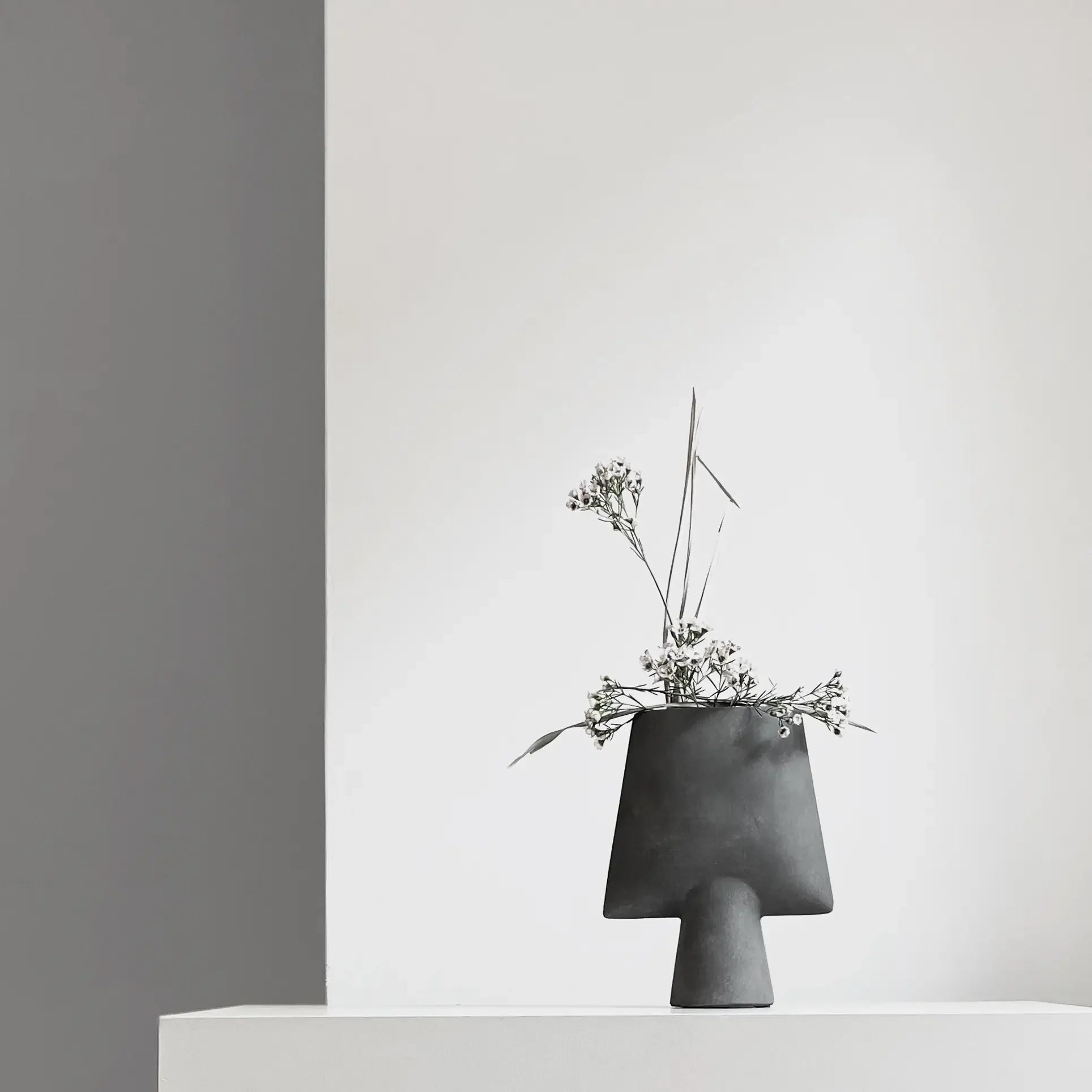 The 101 Copenhagen Sphere Vase Square, Mini—a minimalist black vase—holds white flowers and greenery on a white surface, set against white and gray walls for a sculptural look inspired by ancient Chinese vases.