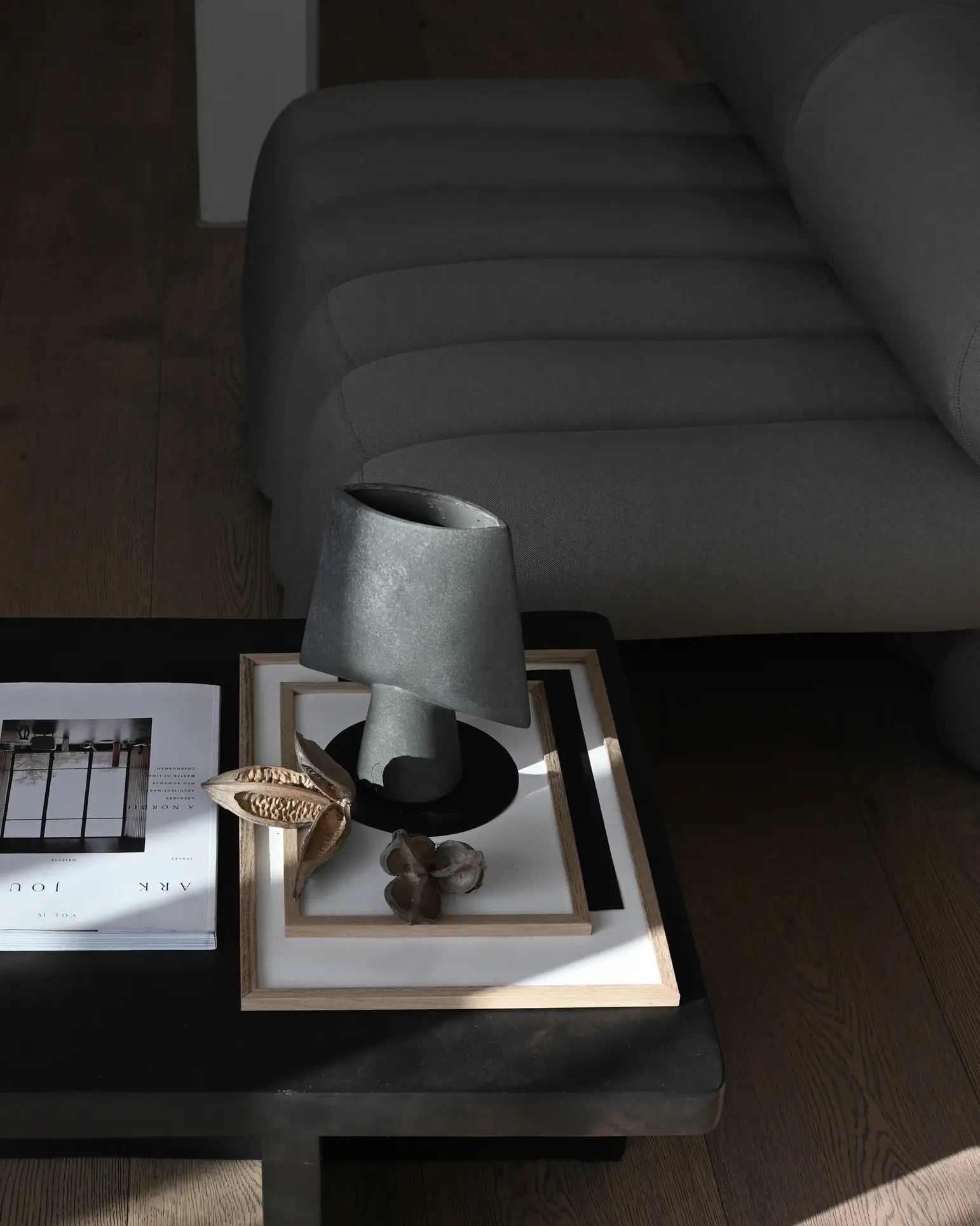 A modern coffee table features the 101 Copenhagen Sphere Vase Square, Mini by 101 Copenhagen, a sculptural gray lamp, a framed tray, and a magazine beside a tufted gray sofa on wooden flooring in natural light.