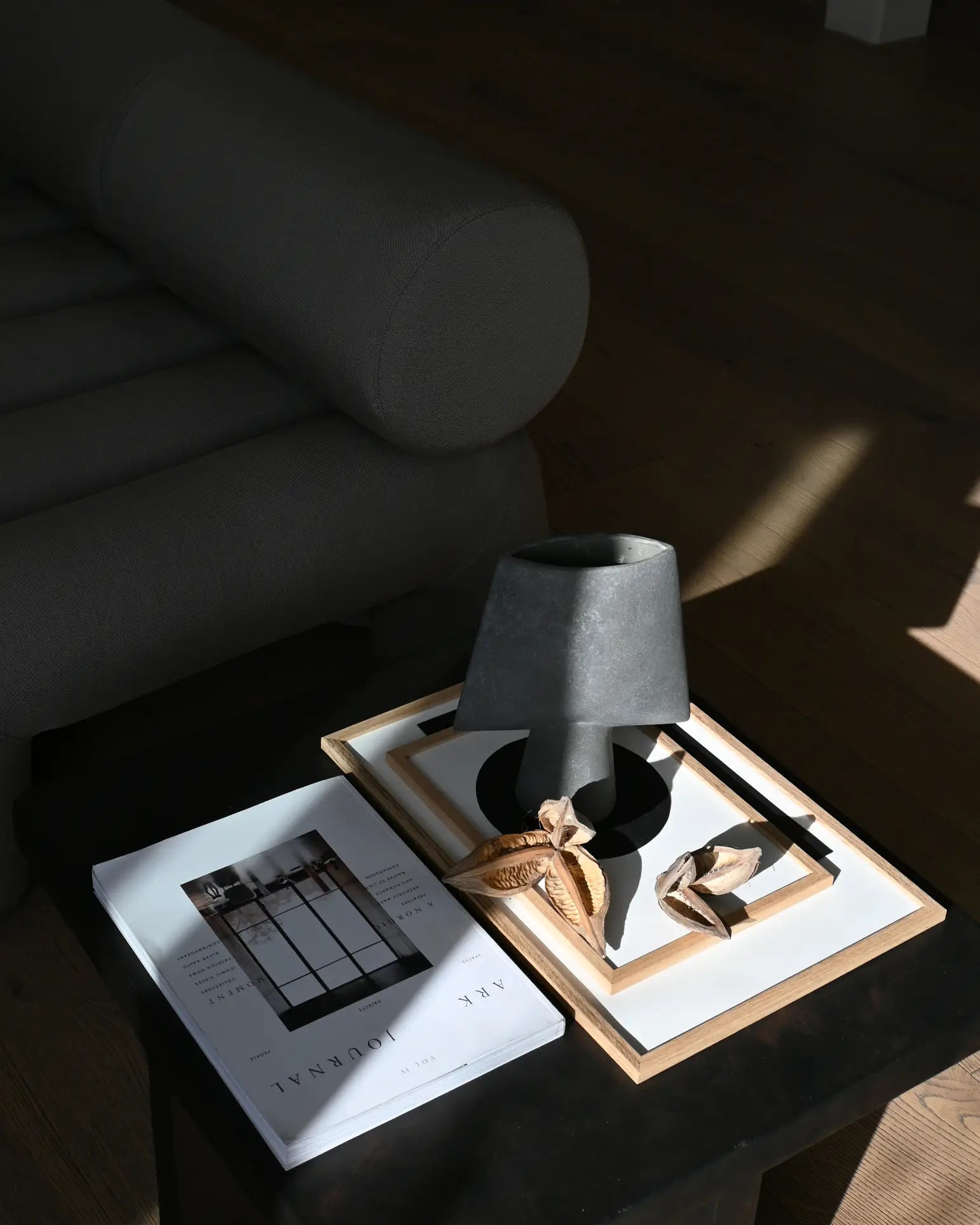 A black side table with sculptural elegance displays a 101 Copenhagen Sphere Vase Square, Mini with dried leaves, a gray lamp, framed art, and the book JAK JOURNAL beside a dark upholstered sofa in a sunlit room with wooden flooring.