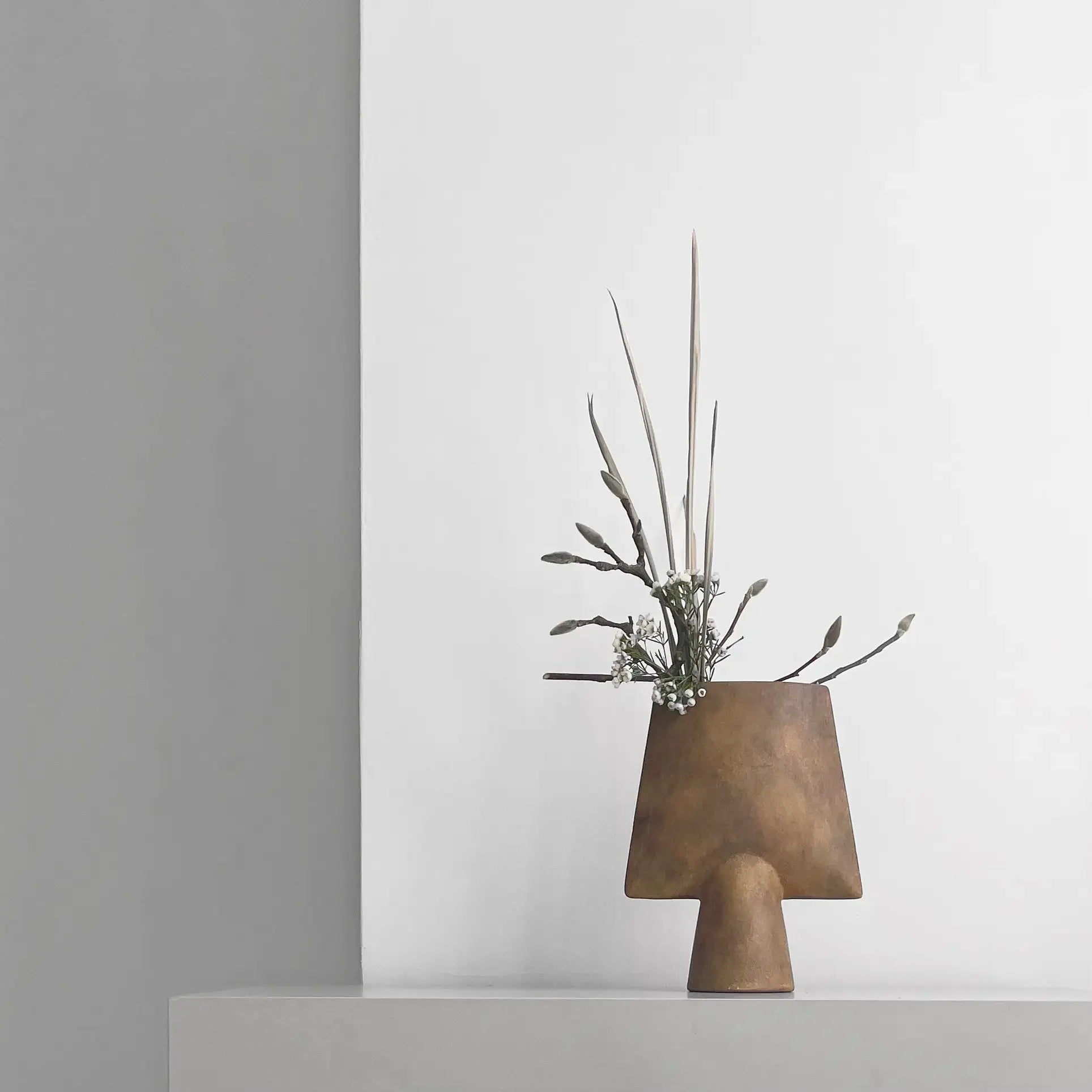The 101 Copenhagen Sphere Vase Square, Mini—a minimalist brown vase—displays sculptural elegance with its geometric design, holding greenery and white flowers on a light gray surface against a white and gray background.
