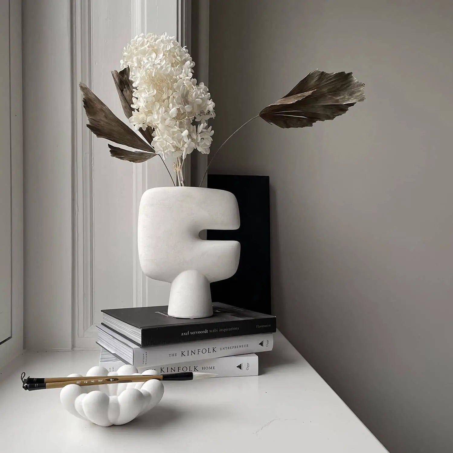 The 101 Copenhagen Tribal Vase, Mini by 101 Copenhagen, styled with dried flowers and palm leaves atop stacked books by a window, forms a minimal monochrome scene alongside a sculptural white ink brush holder.