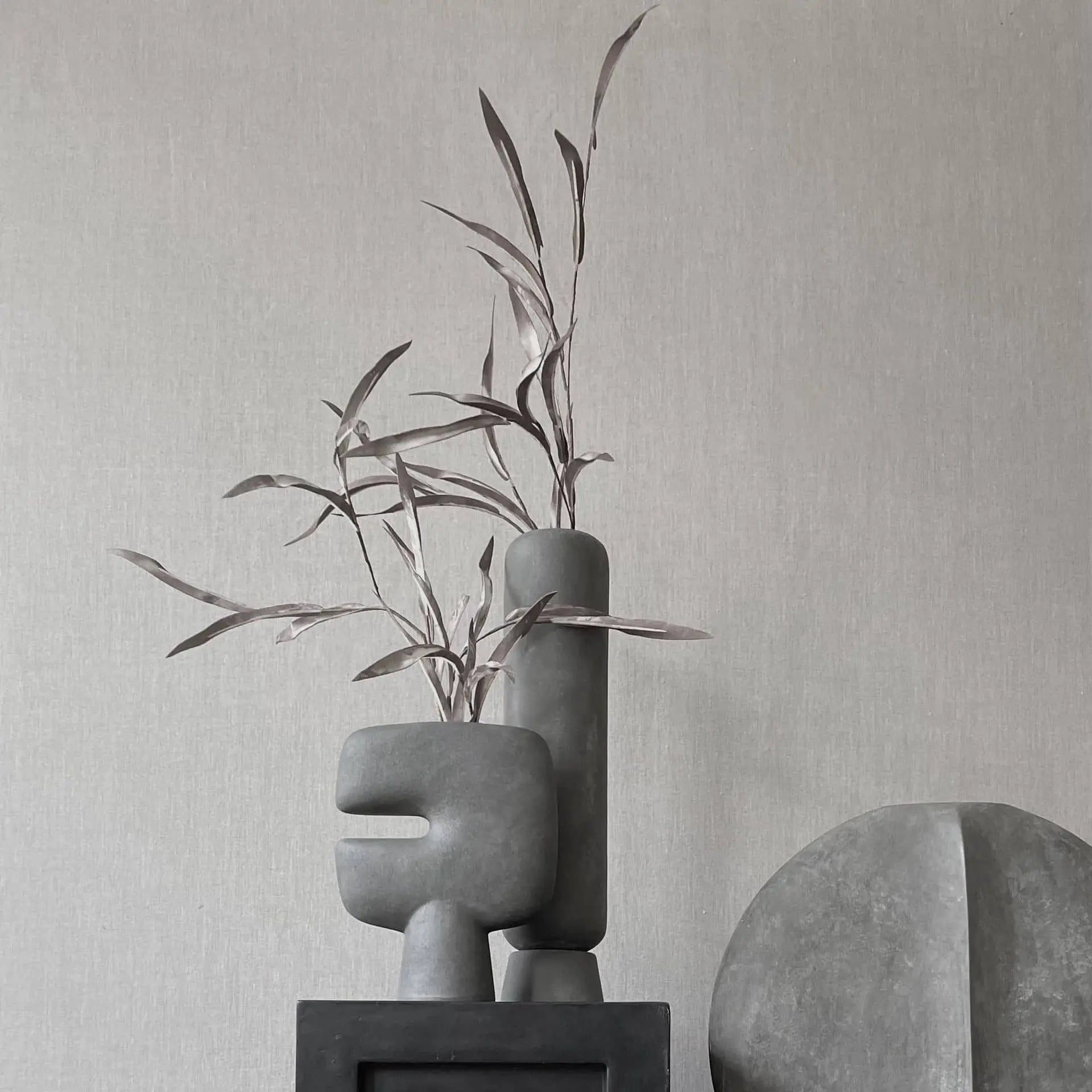 Minimalist composition featuring the 101 Copenhagen Tribal Vase, Mini by 101 Copenhagen—a matte gray sculptural vase—paired with a terracotta vase and round gray sculpture, set against a textured light gray background for a modern, muted style.