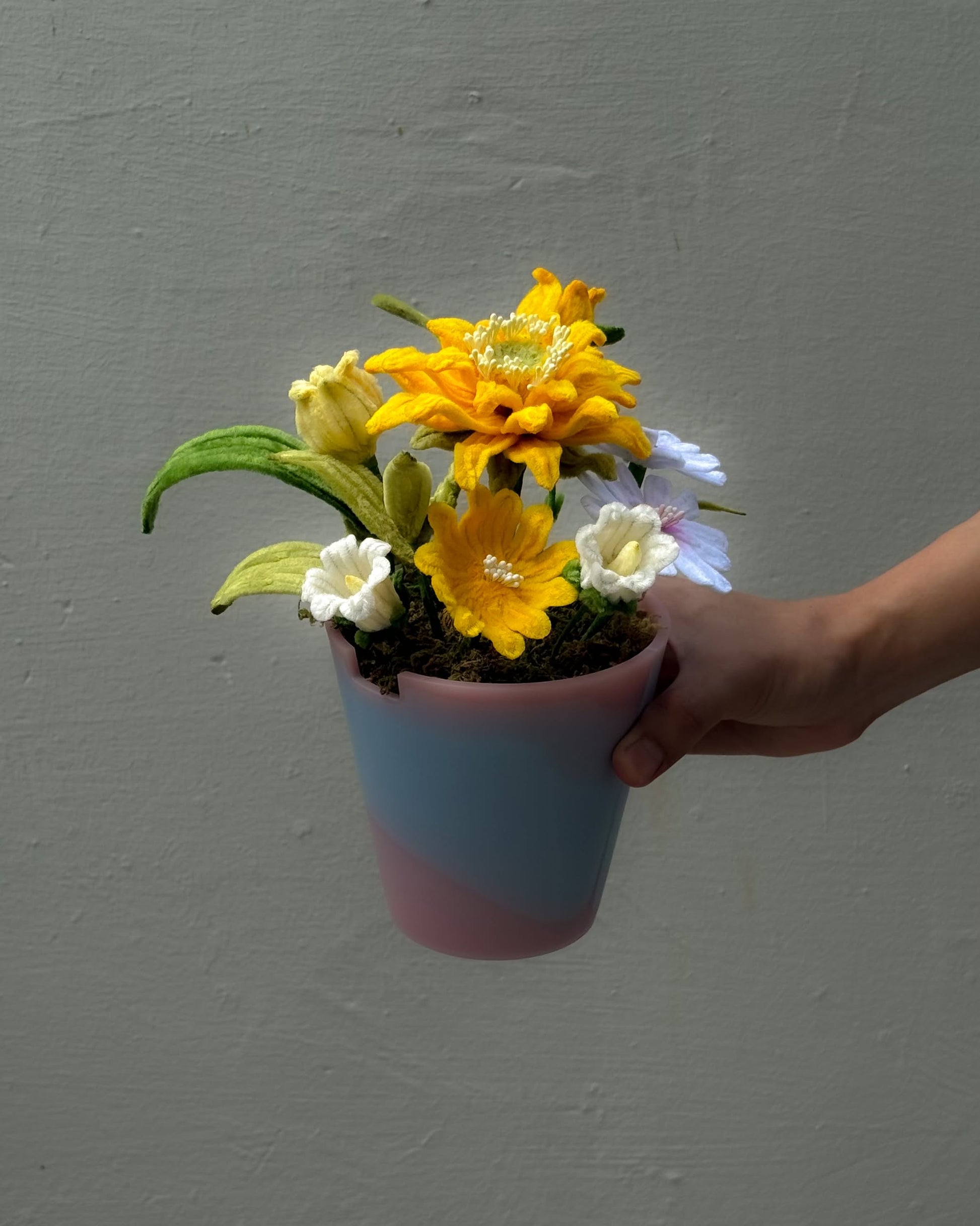 A hand holds the Ah!GUGU x Stacked Store Bloom in Golden Hour, a pastel recycled plastic pot with handcrafted artificial yellow, white, and light purple flowers by Ah!GUGU, set against a plain gray background.
