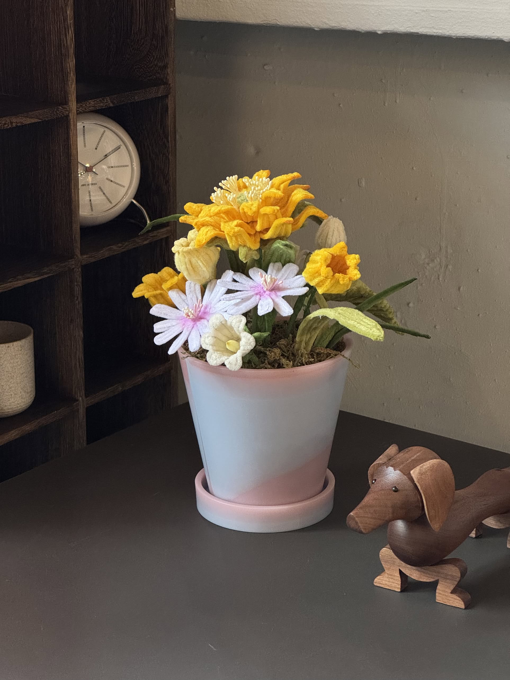 The Ah!GUGU x Stacked Store Bloom in Golden Hour - Handcrafted Flowers sits in a recycled plastic pot beside a wooden dog figurine, with a wooden cubby and white clock seen in the background.
