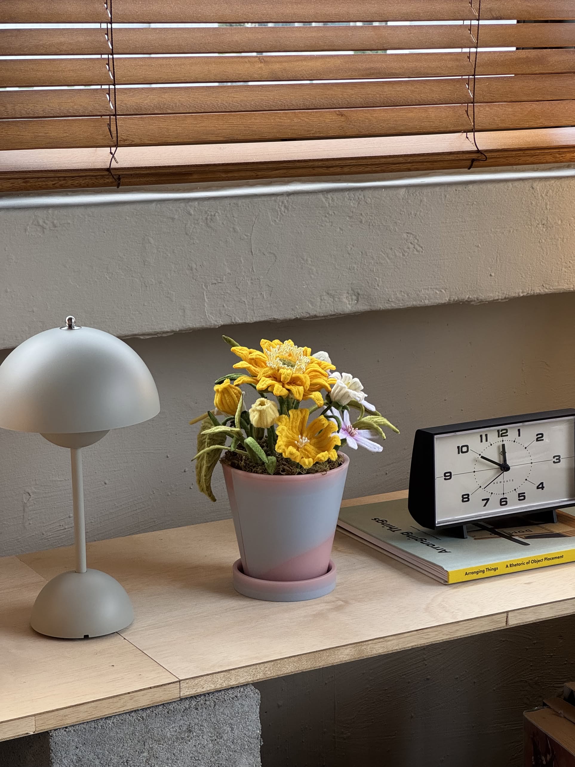 A small desk with a modern gray lamp, Ah!GUGU x Stacked Store Bloom in Golden Hour handcrafted flowers in a recycled pot with yellow and white blooms, a black and white clock, set against beige walls and window blinds.