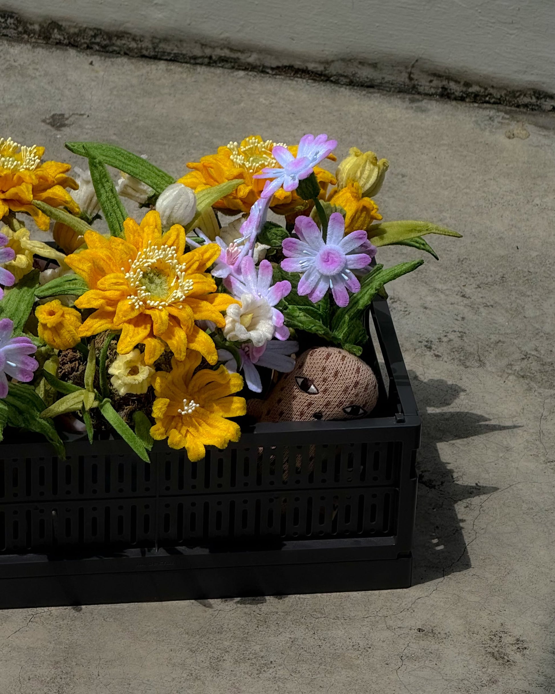 A black recycled plastic pot from Ah!GUGU holds the Ah!GUGU x Stacked Store Bloom in Golden Hour – Handcrafted Flowers, featuring artificial yellow, white, and light purple blooms. A painted rock with a face peeks out among the florals.