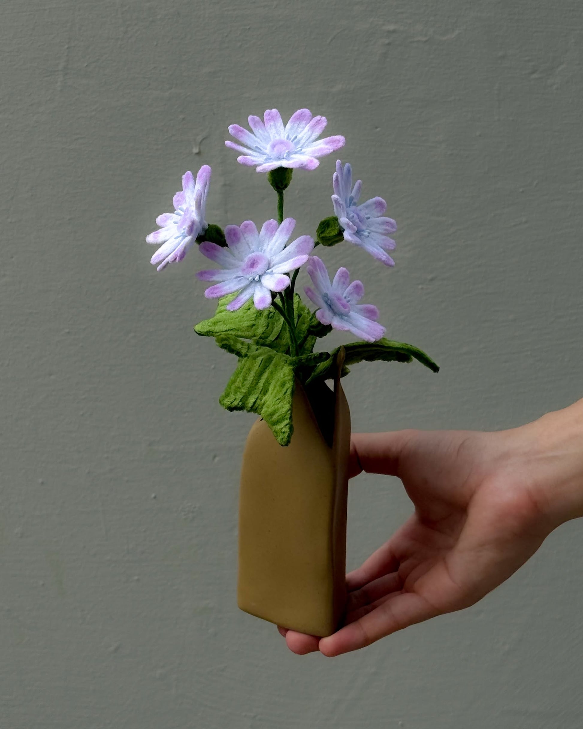 A hand holds the Ah!GUGU x Stacked Store Still Life in Bloom - Handcrafted Flowers, featuring delicate light purple fabric flowers with green leaves in a sculptural vase, set against a plain gray background.