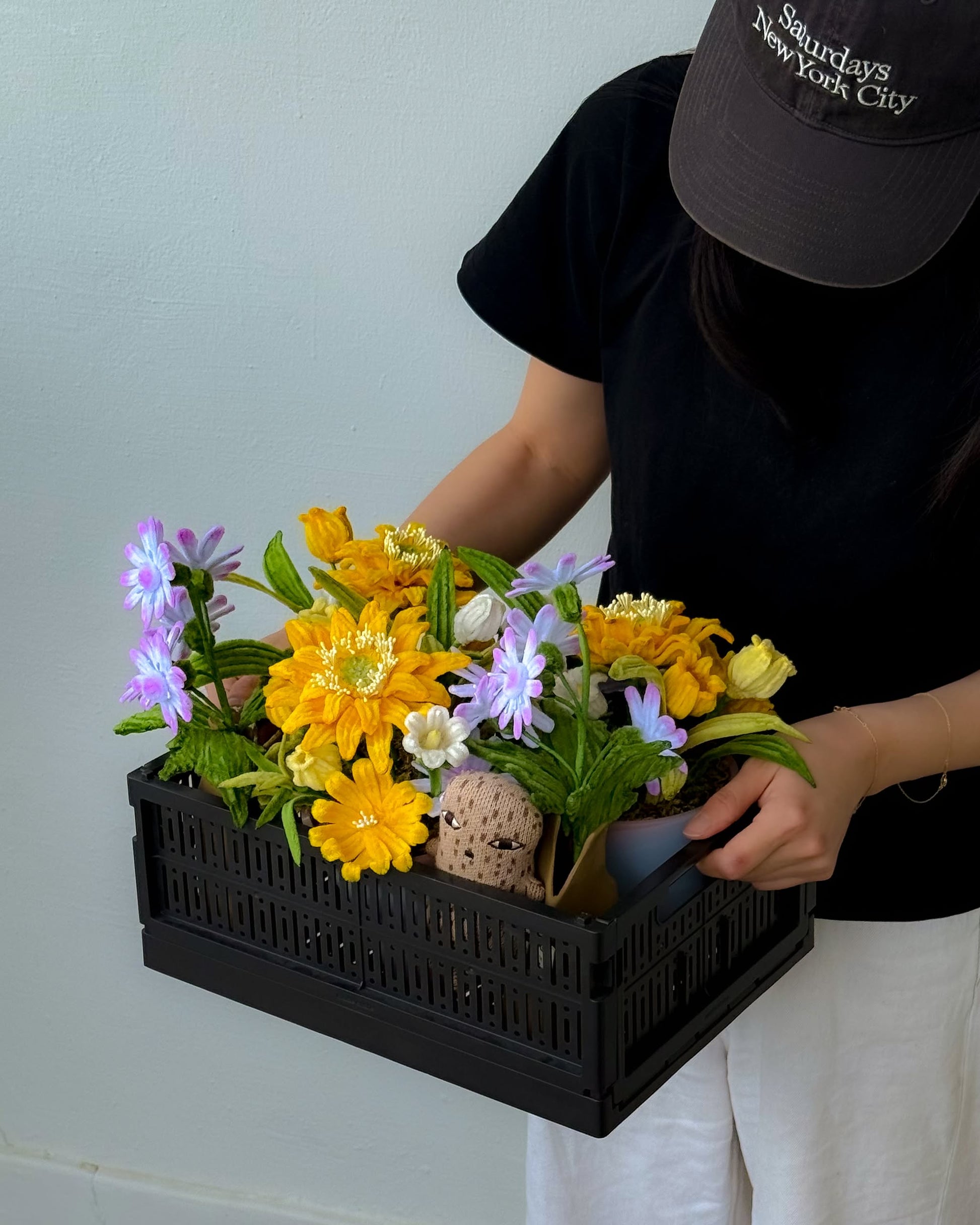 A person in a black shirt and cap holds the Ah!GUGU x Stacked Store Still Life in Bloom - Handcrafted Flowers, featuring vibrant artificial yellow and purple blooms, with a plain light-colored wall as the backdrop.