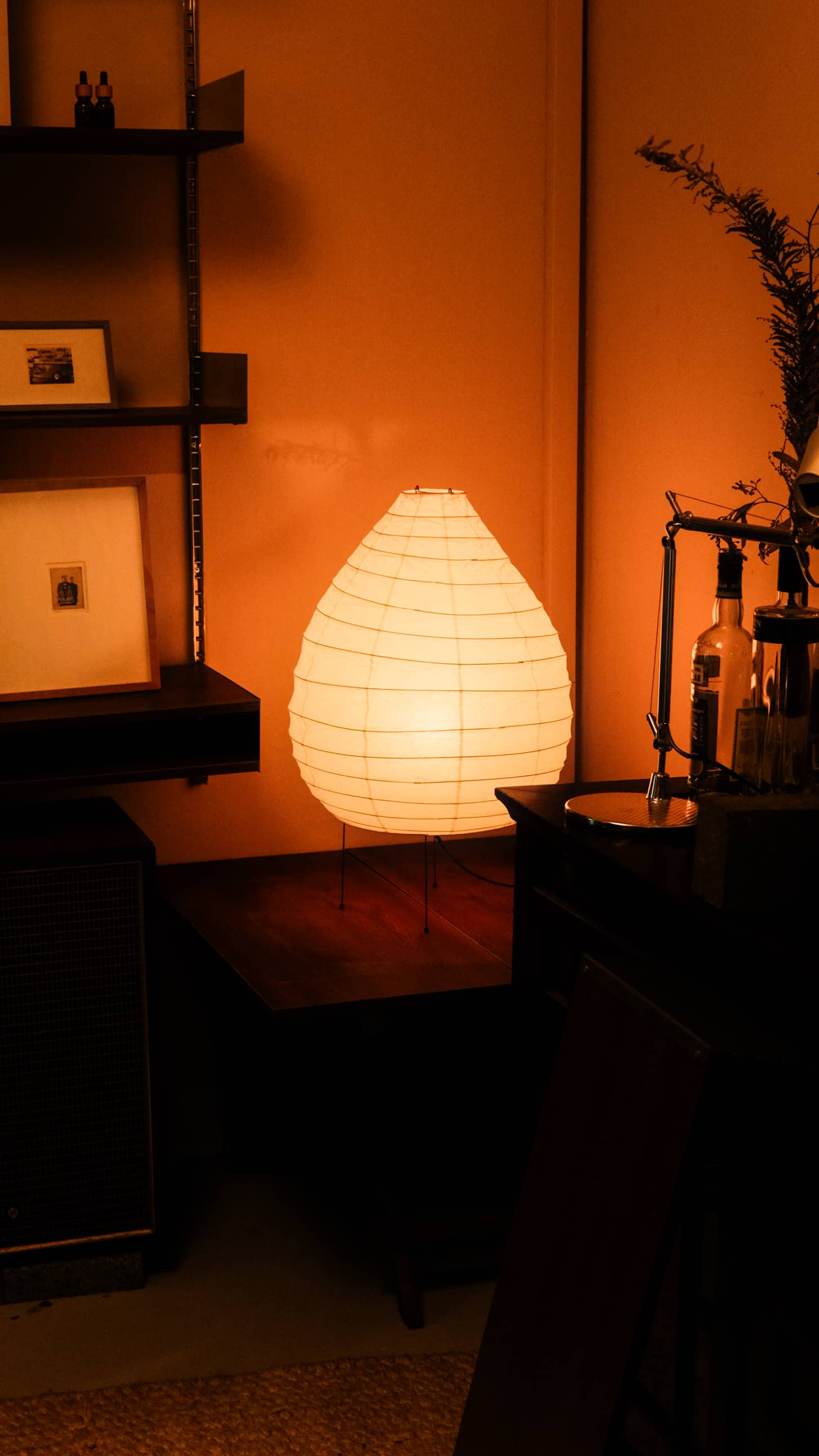 The Akari 1N paper lantern lamp by Akari casts a warm glow on a wooden table in a dimly lit room, surrounded by shelves, framed art, and decorative items.