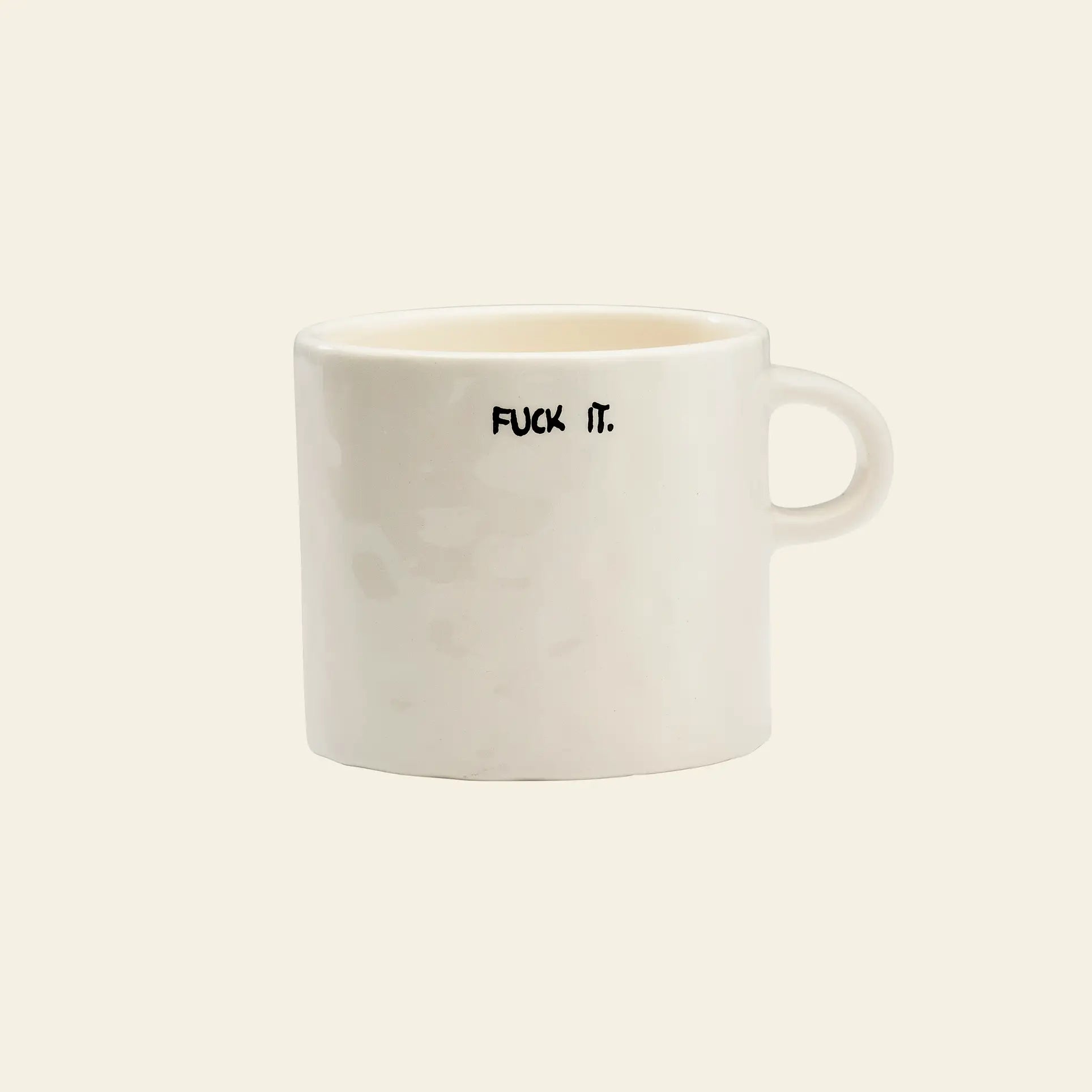 The Anna + Nina Fuck It Mug by Anna + Nina is a plain white ceramic mug with a handle, featuring “FUCK IT.” in bold black uppercase letters on one side—perfect for adding attitude to your coffee or tea break. Set against a light beige background.