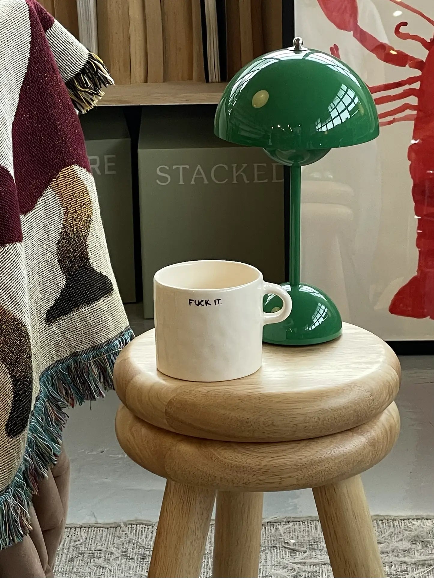 The Anna + Nina Fuck It Mug, featuring fuck it. on the front, rests on a wooden stool beside a green lamp. A fringed blanket and lobster artwork in the background add cozy, playful touches—perfect for coffee breaks.