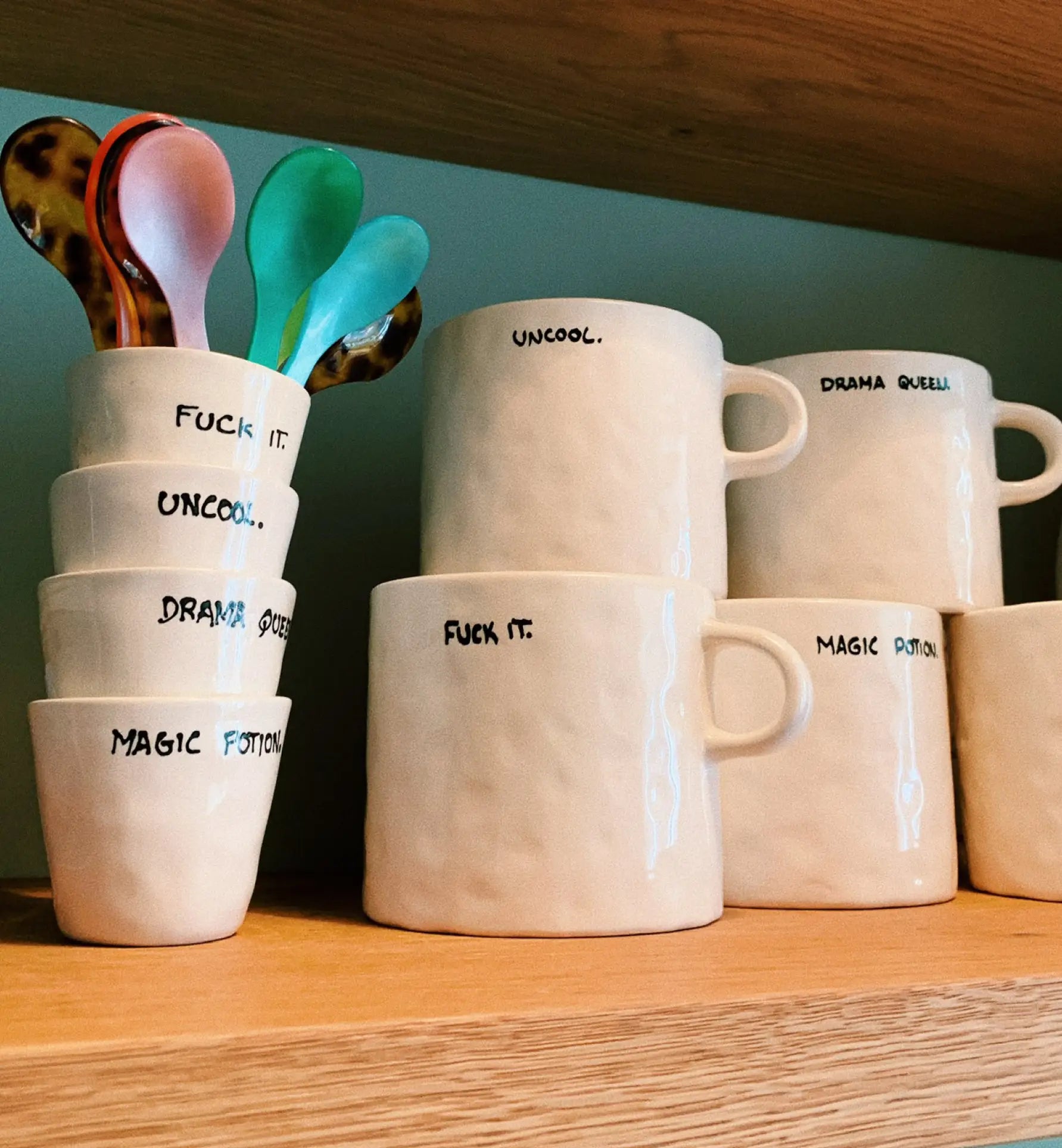 The Anna + Nina Fuck It Mug, a cream-colored ceramic mug with playful handwritten text, sits on a wooden shelf alongside coordinating cups and colorful spoons—perfect for brightening up your coffee or tea break.