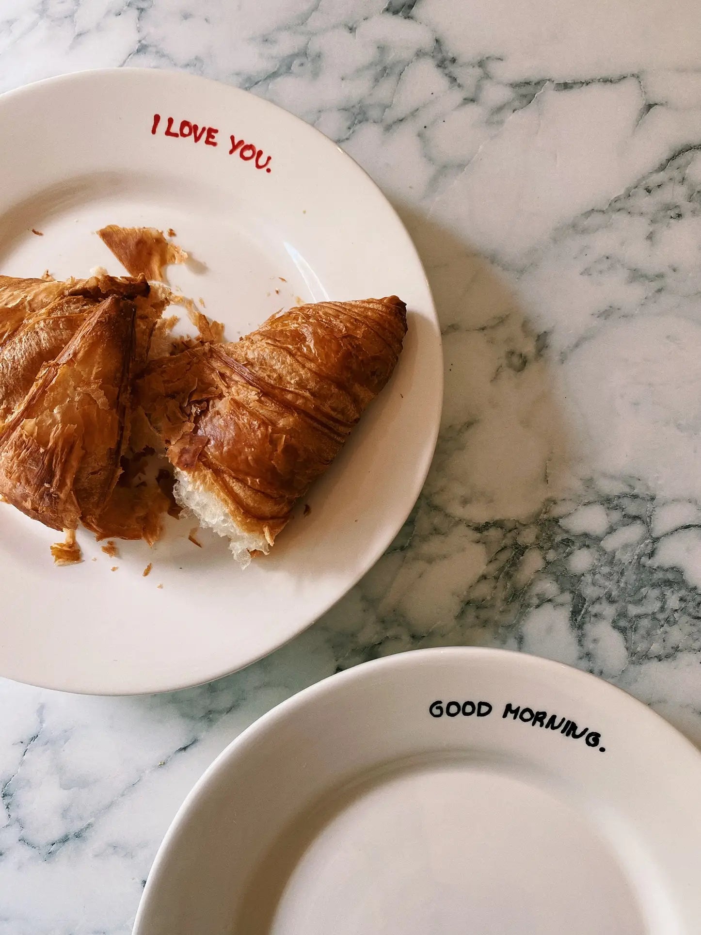 A croissant, broken in half, sits on a plate labeled “I LOVE YOU” beside an empty Anna + Nina Good Morning Breakfast Plate—adding a cheerful touch from Anna + Nina to your breakfast routine atop a marble surface.