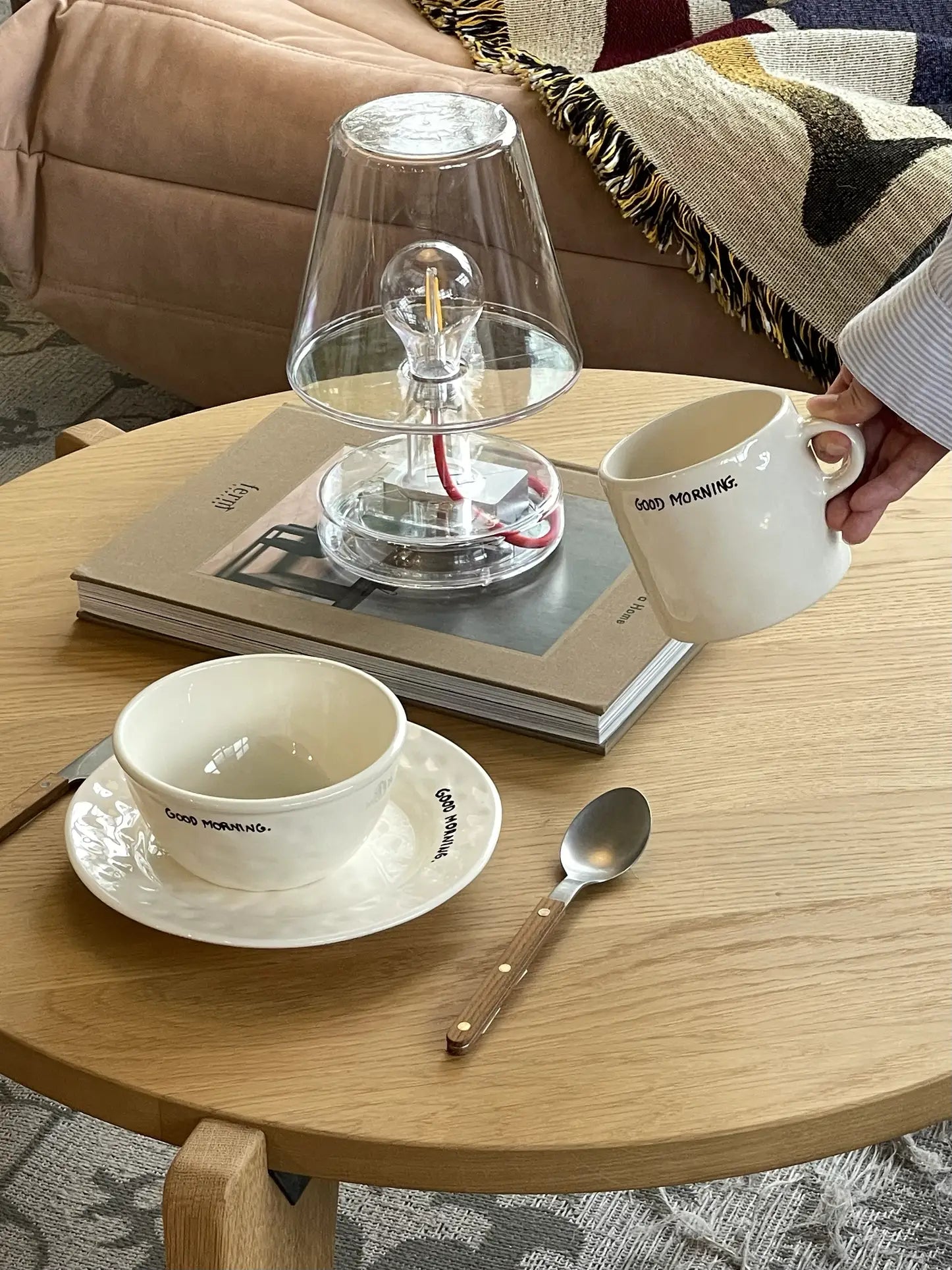 A hand holds the Anna + Nina Good Morning Mug near a wooden coffee table with a matching bowl and saucer, spoon, glass lamp, and books. In the background, a sofa with a patterned blanket enhances the cozy atmosphere.