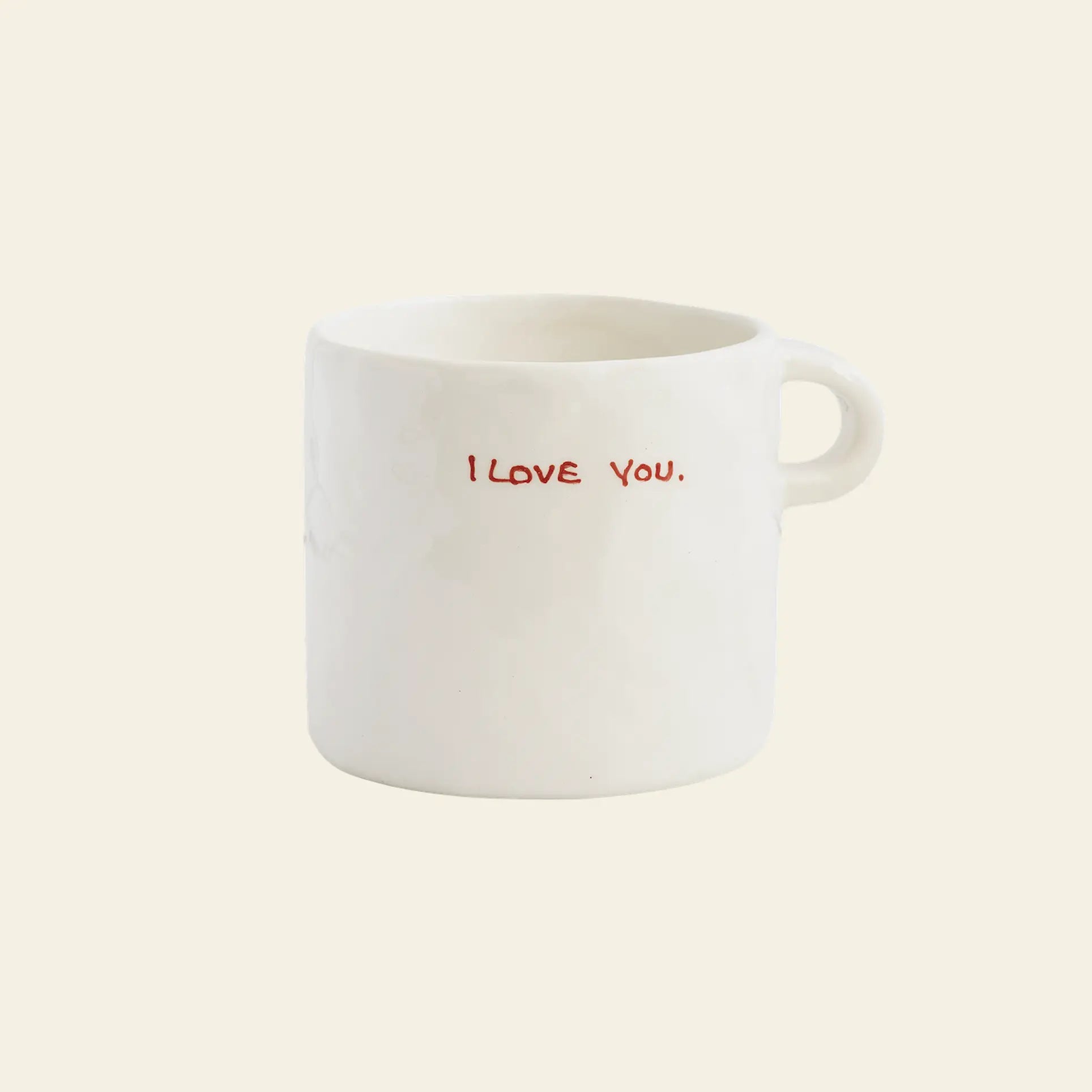The Anna + Nina I Love You Mug by Anna + Nina is a white ceramic mug with a small handle and I LOVE YOU. in red capitals on the front, perfect for showing affection during your coffee. Plain beige background.