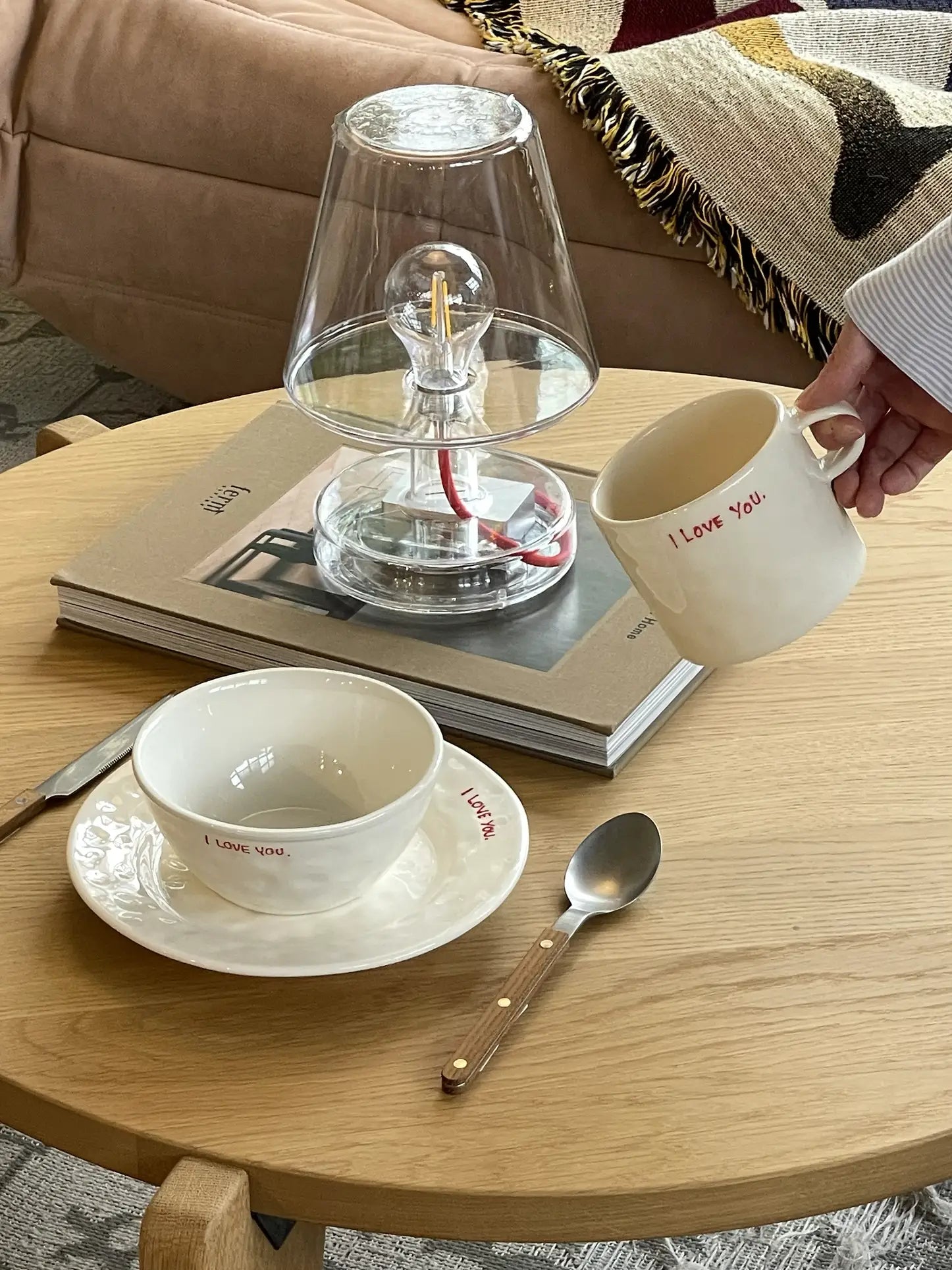 A hand holds the Anna + Nina I Love You Mug—ideal for morning coffee and sharing affection. On the table: a matching Anna + Nina bowl and plate, spoon, two books, a glass lamp. A beige sofa with a patterned blanket is in the background.