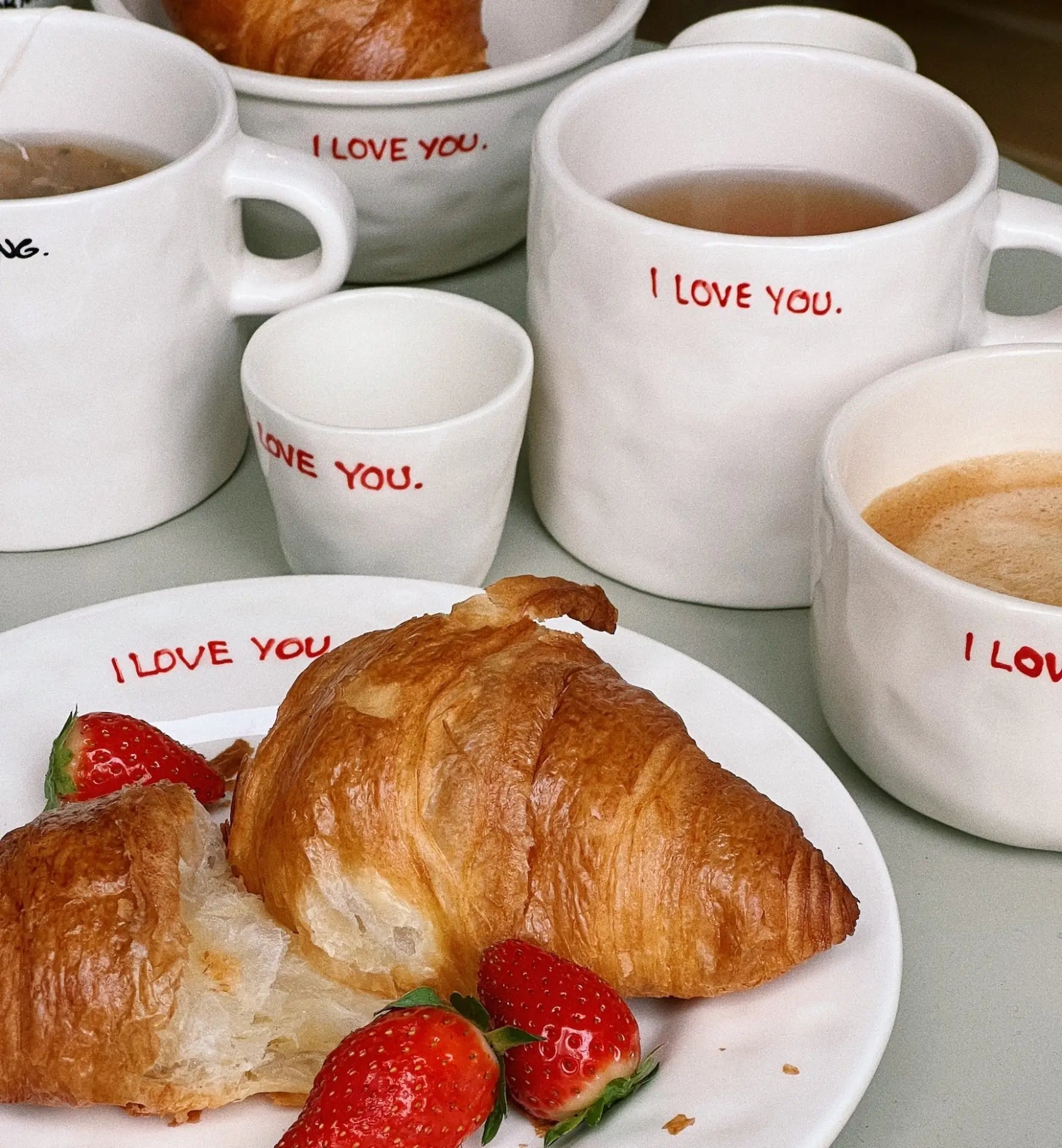 A cozy breakfast scene featuring croissants and strawberries, served with morning coffee and tea in Anna + Nina I Love You Mugs. The red text on the mugs adds a heartfelt touch, perfect for expressing affection.