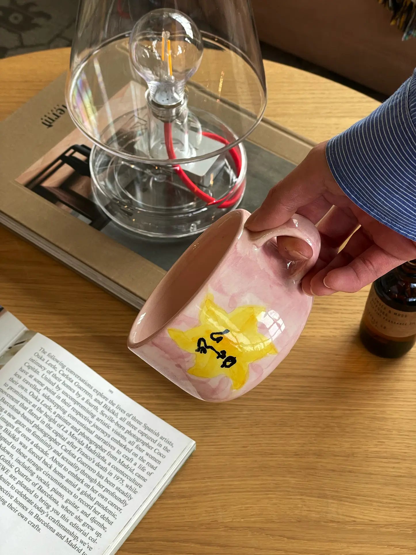 A hand holding the Anna + Nina Sunny Side Up Cappuccino Mug, featuring a yellow star and black writing, above a wooden table with a book, magazine, clear lamp, and bottle—perfect for a cozy coffee moment.