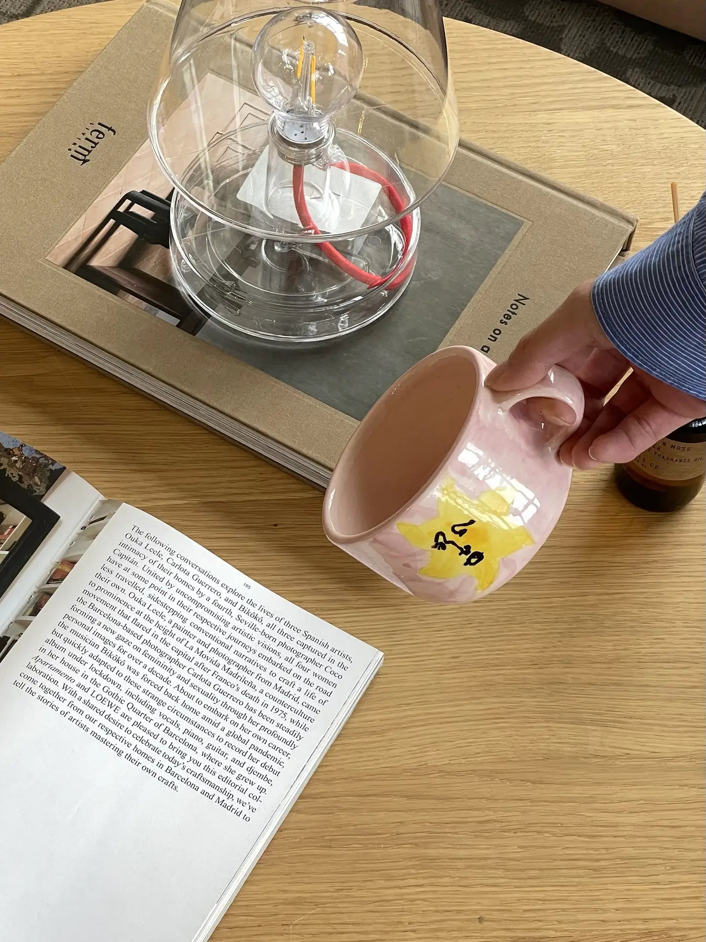 A person in a blue shirt holds the Anna + Nina Sunny Side Up Cappuccino Mug over a wooden table with an open book, closed book, glass coffee maker, and small brown bottle.