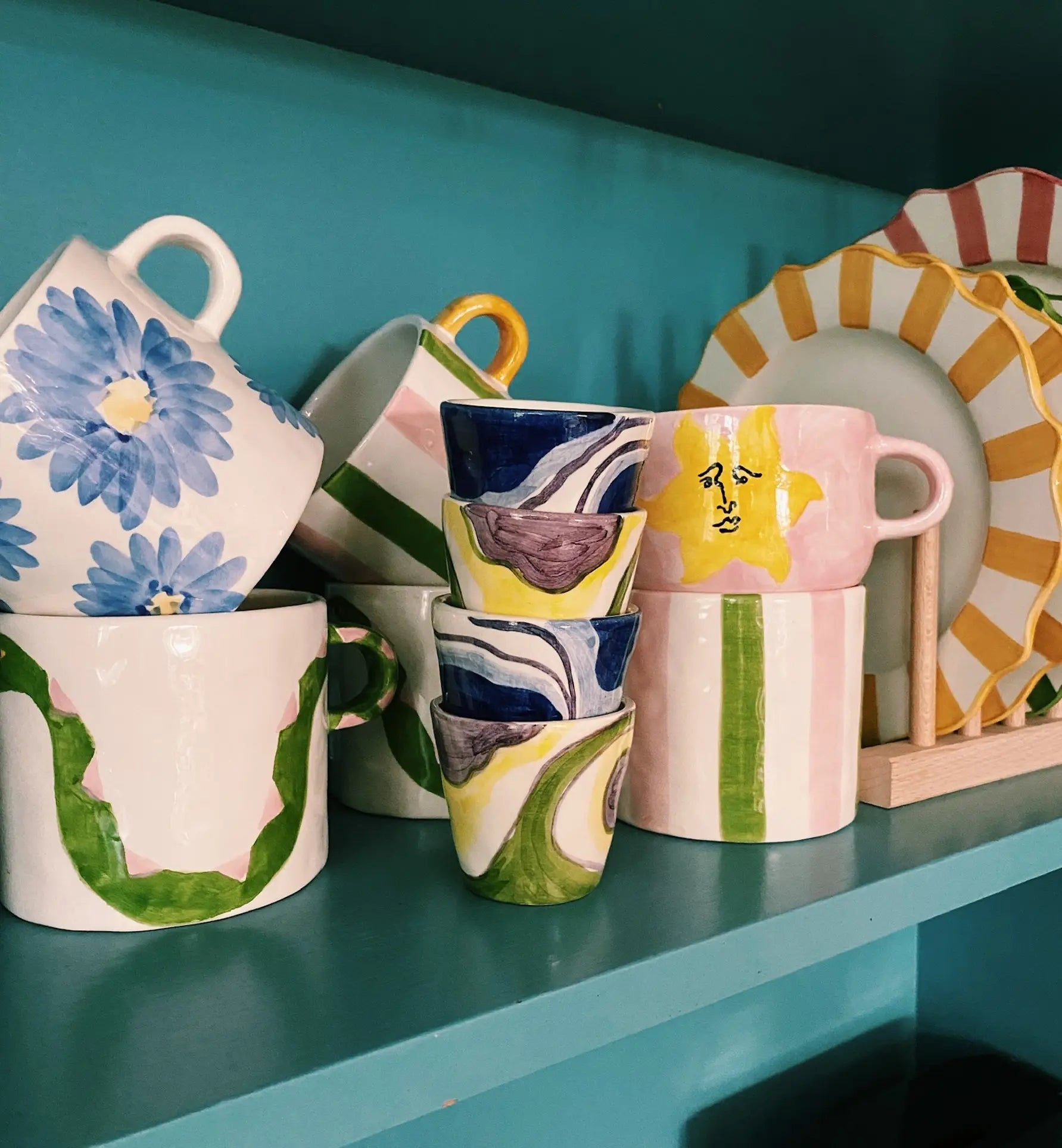 A vibrant display of Anna + Nina ceramics, including the Sunny Side Up Cappuccino Mug, sits on a teal shelf. The hand-painted mugs and plates show floral, abstract, and striped designs in green, blue, yellow, pink, and white.