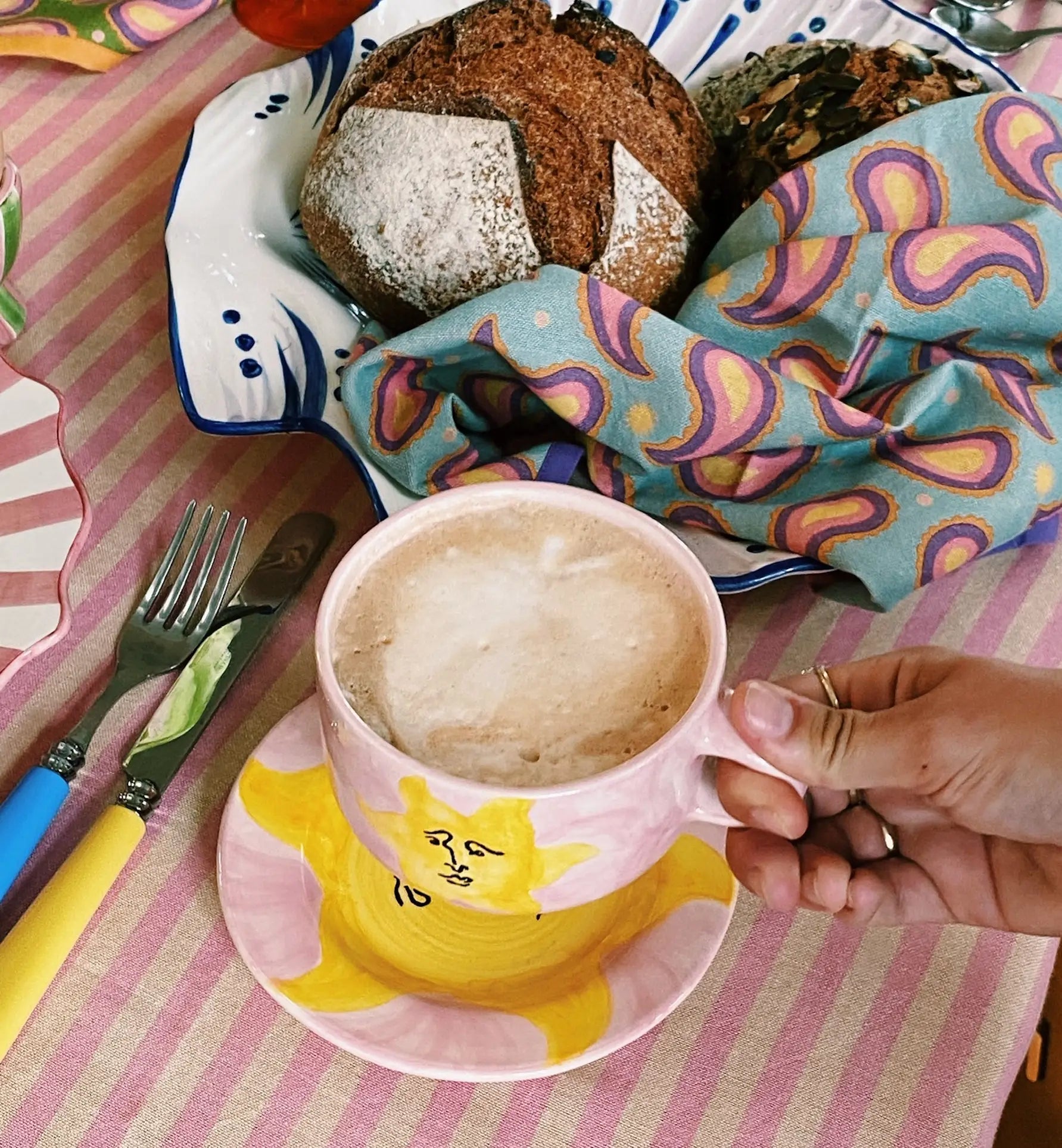 A hand holds the Anna + Nina Sunny Side Up Cappuccino Mug, filled with foamy coffee and set on a matching saucer. A knife, fork, and two loaves of bread rest on a pink striped tablecloth for a cheerful coffee moment.